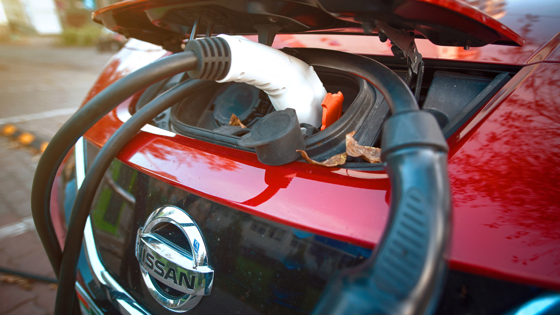 A close-up of a white charging cable plugged into the front of a red Nissan car.