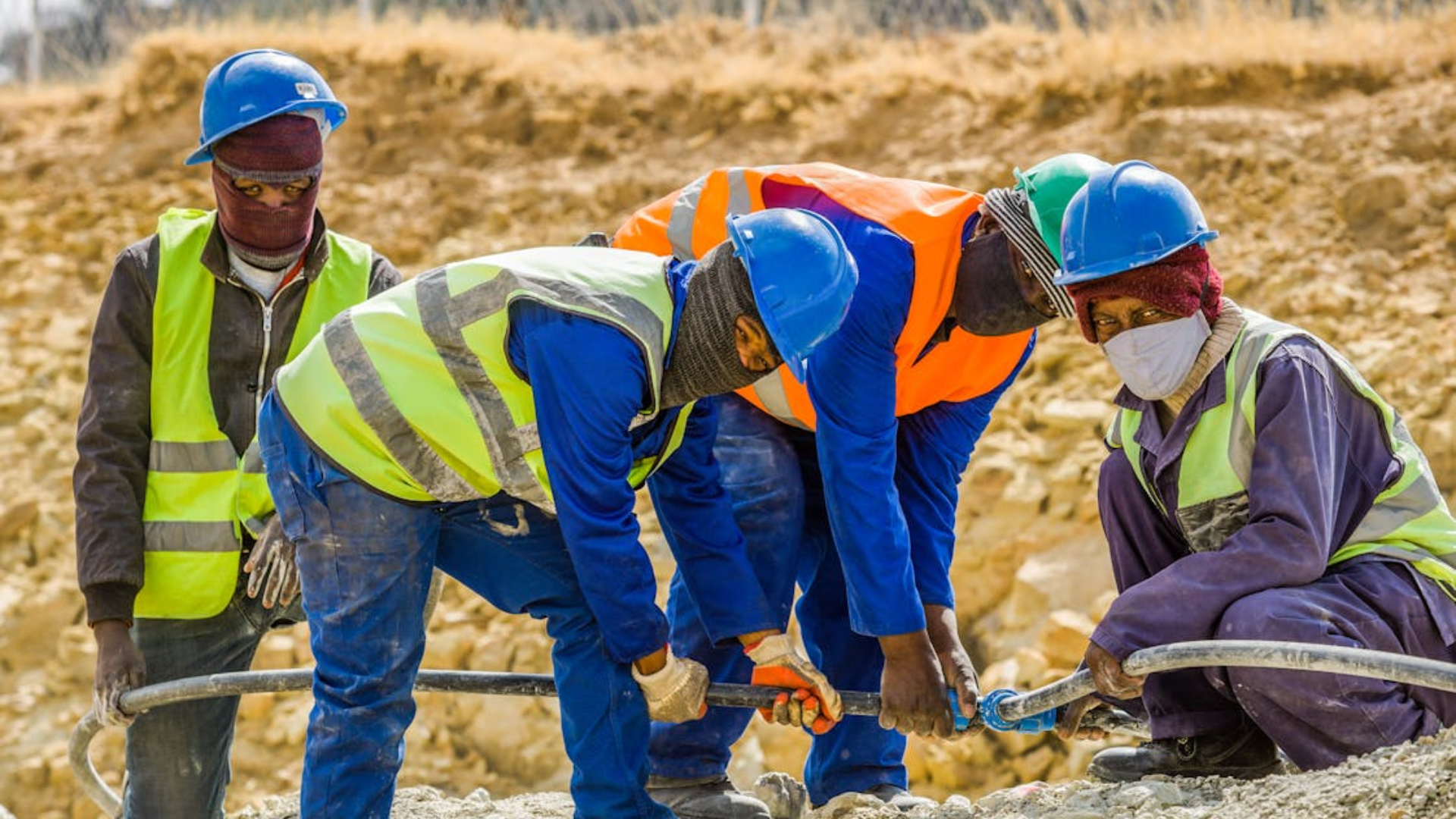 Four construction workers wearing hard hats and safety vests pull a long cable on a rocky dirt site.