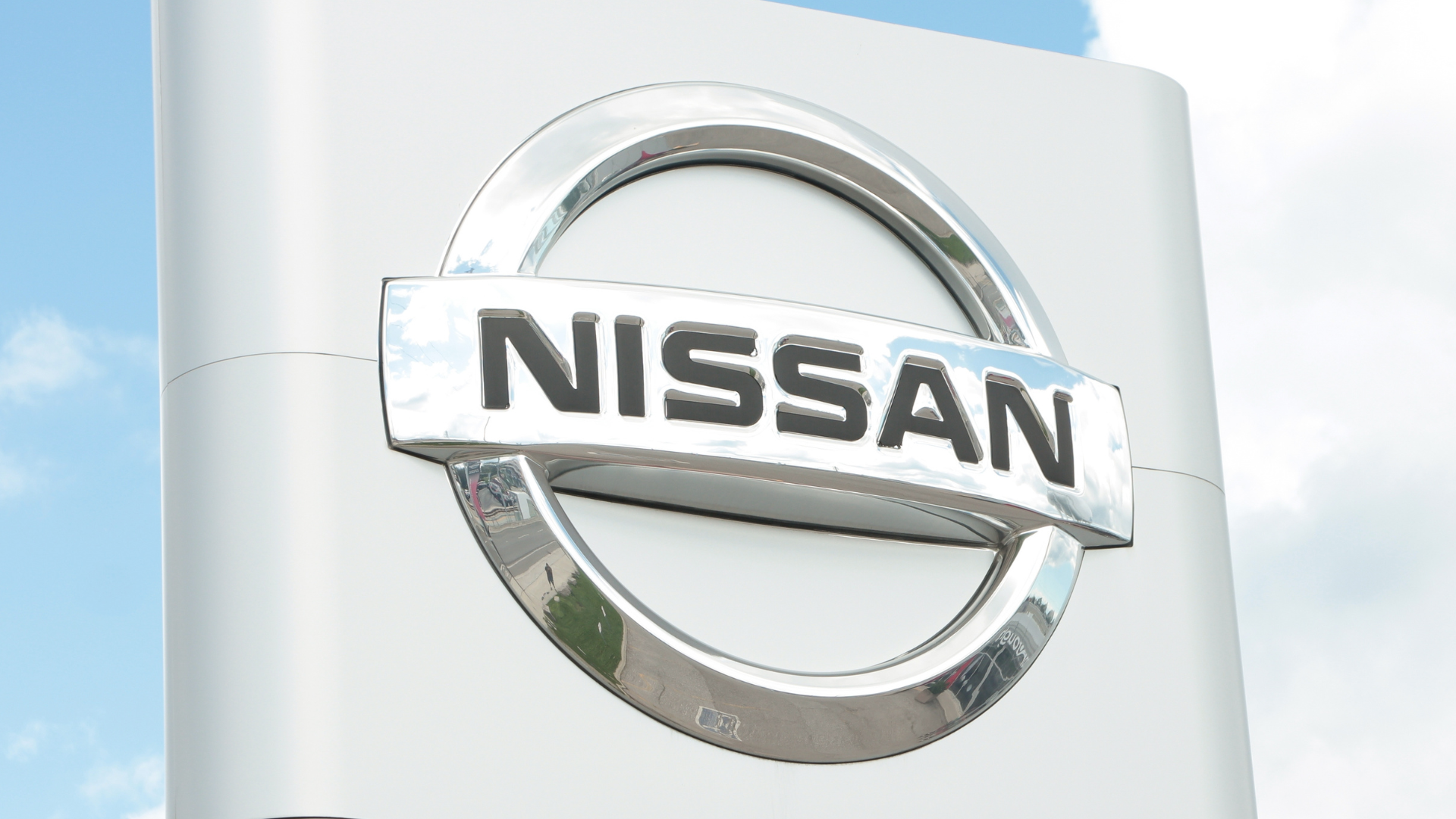 A large, shiny silver Nissan logo sign stands against a light blue sky.