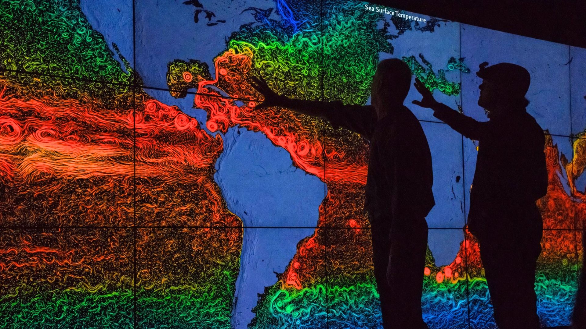 Two people pointing at a global sea surface temperature map display showing swirling ocean current patterns.