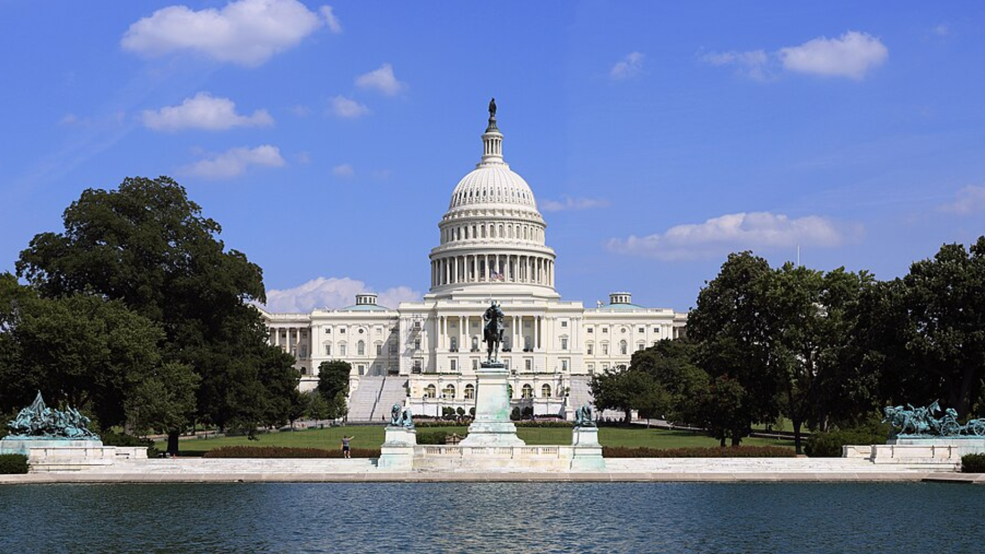  A front-on shot of the United States Capitol Building with its large white dome under a clear blue sky, seen across a reflecting pool.
