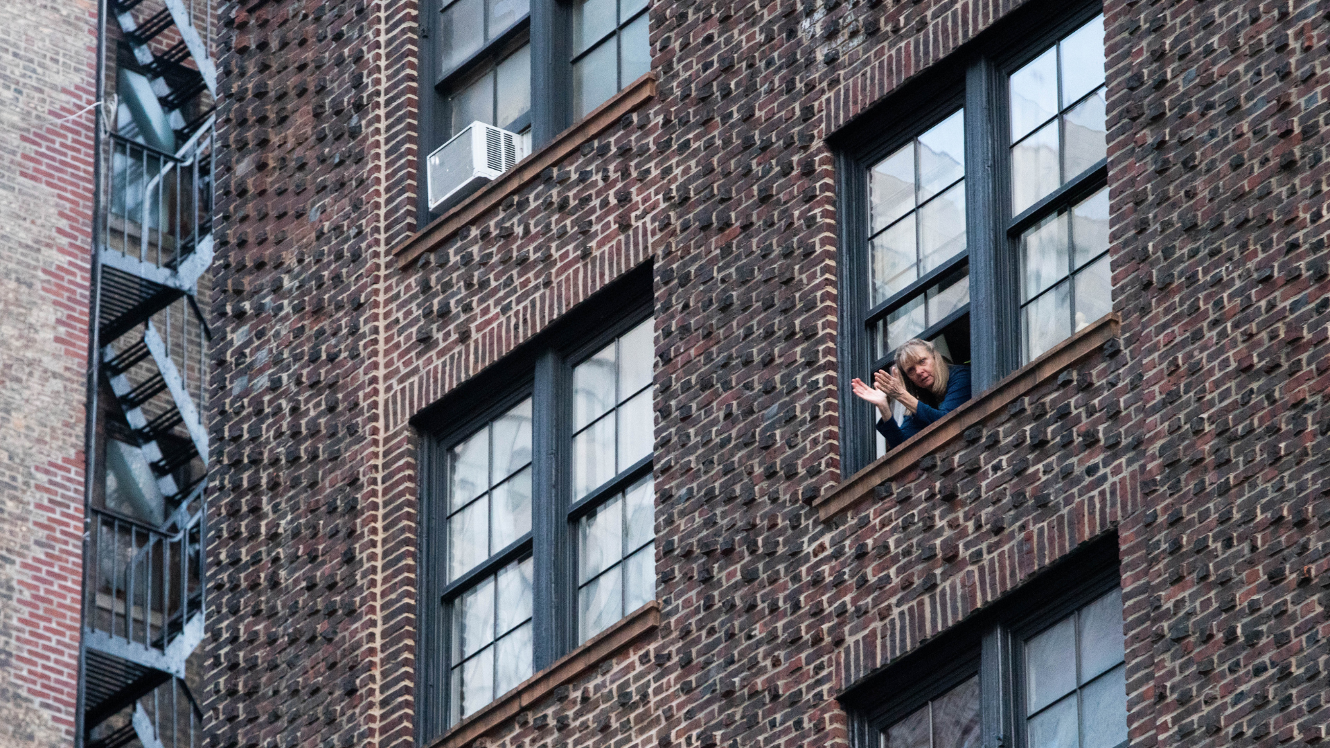 A person leans out of an apartment window in a brick building and claps their hands.