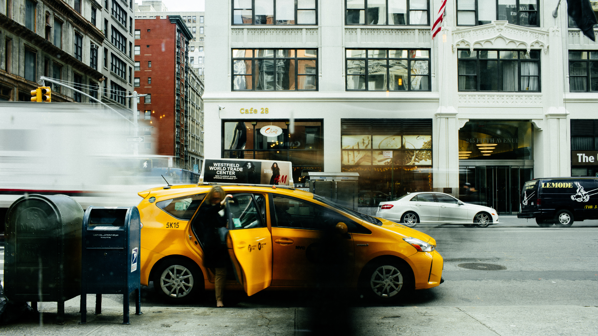 A person steps out of a bright yellow taxi parked on a city street next to blue and green mailboxes