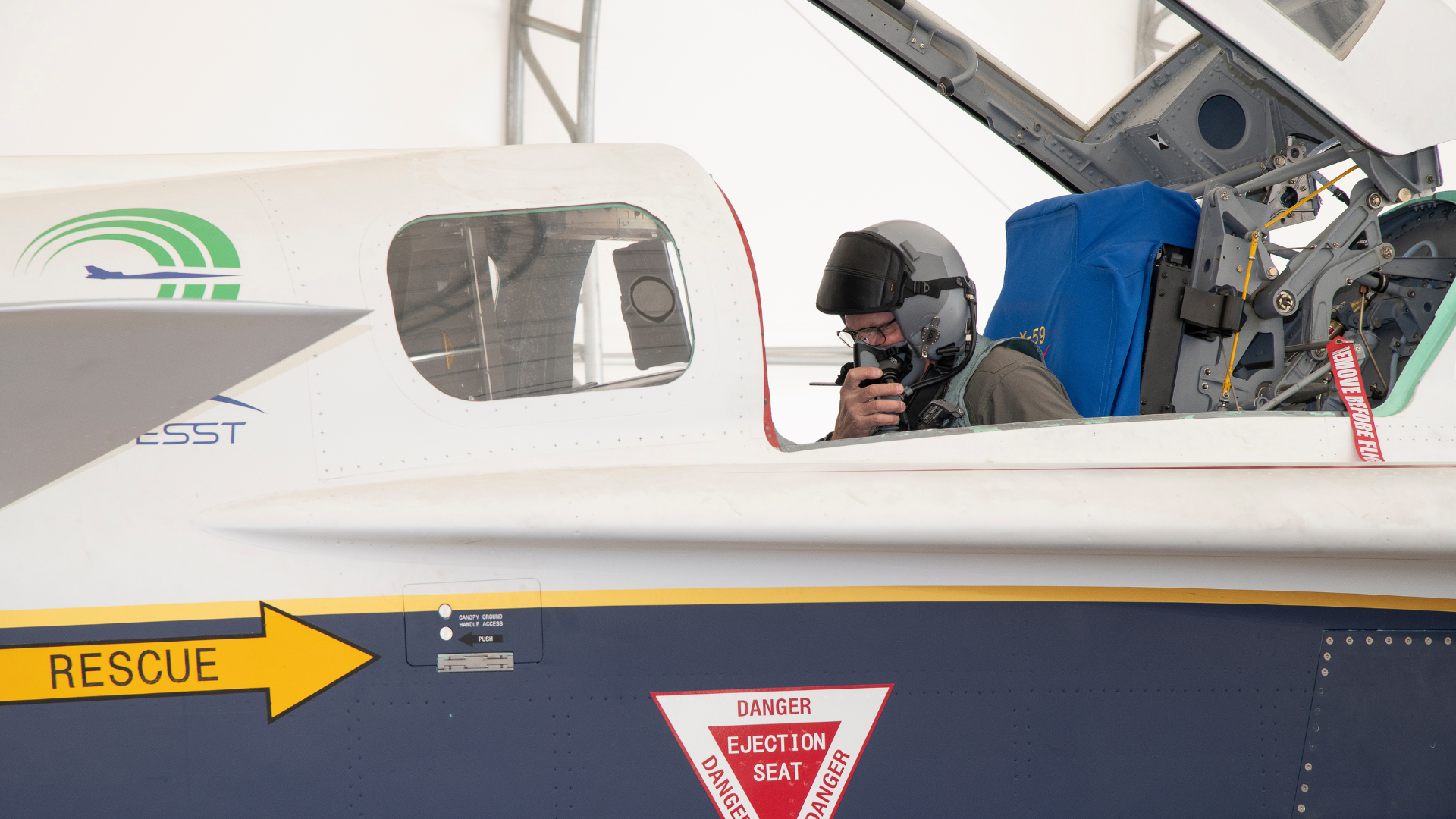 A test pilot wearing a flight helmet and oxygen mask sits in the open cockpit of the X-59 aircraft during pre-flight checks.