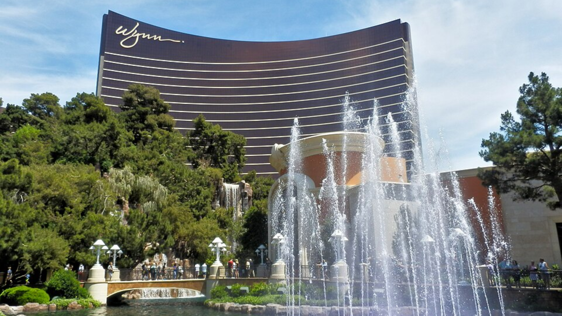  A wide view of the Wynn Las Vegas resort featuring an outdoor pond with dancing fountains and lush landscaping in the foreground.