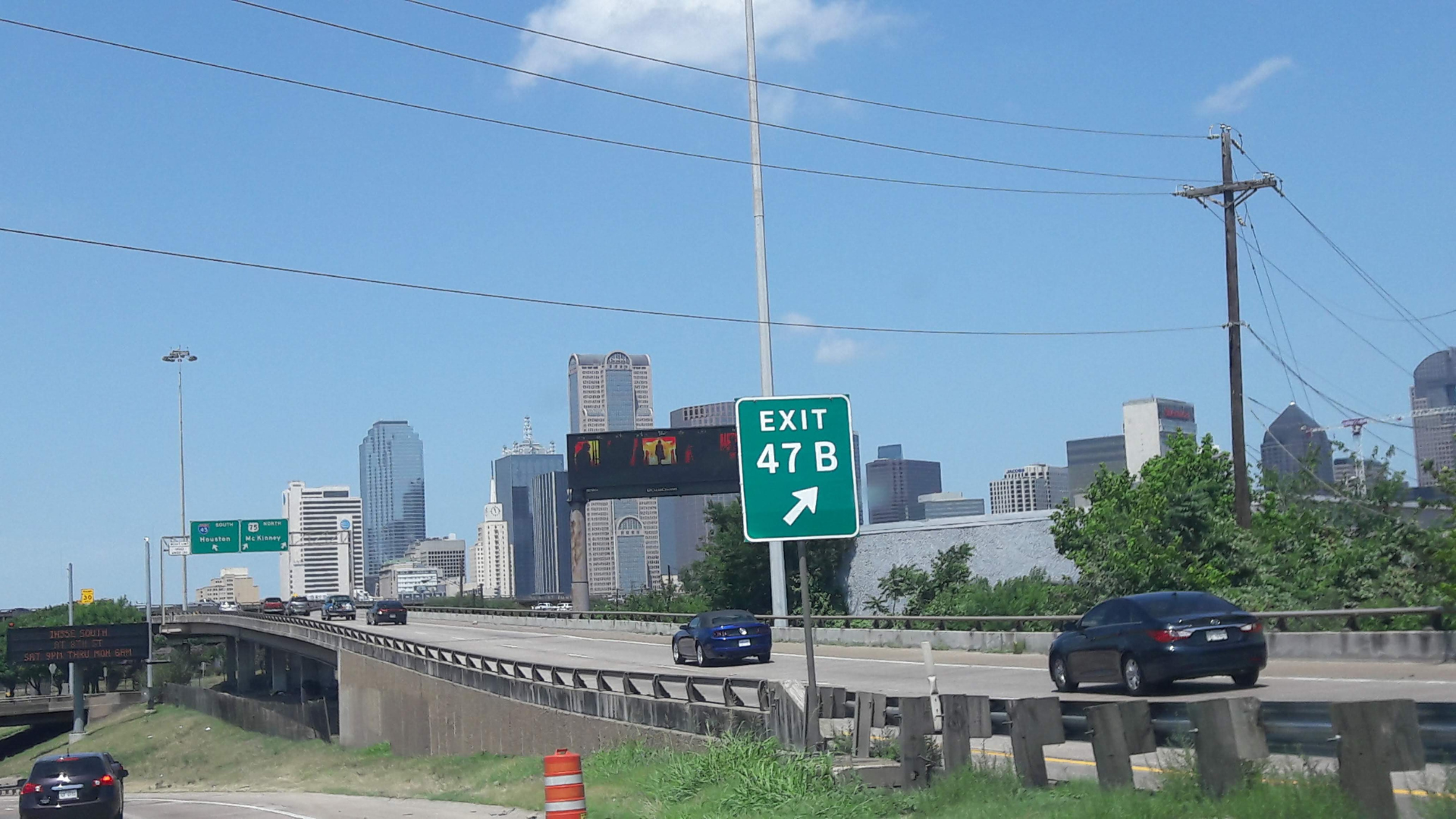 Cars drive along a multi-lane highway toward the Dallas city skyline, with a prominent green _EXIT 47 B_ sign in the foreground.