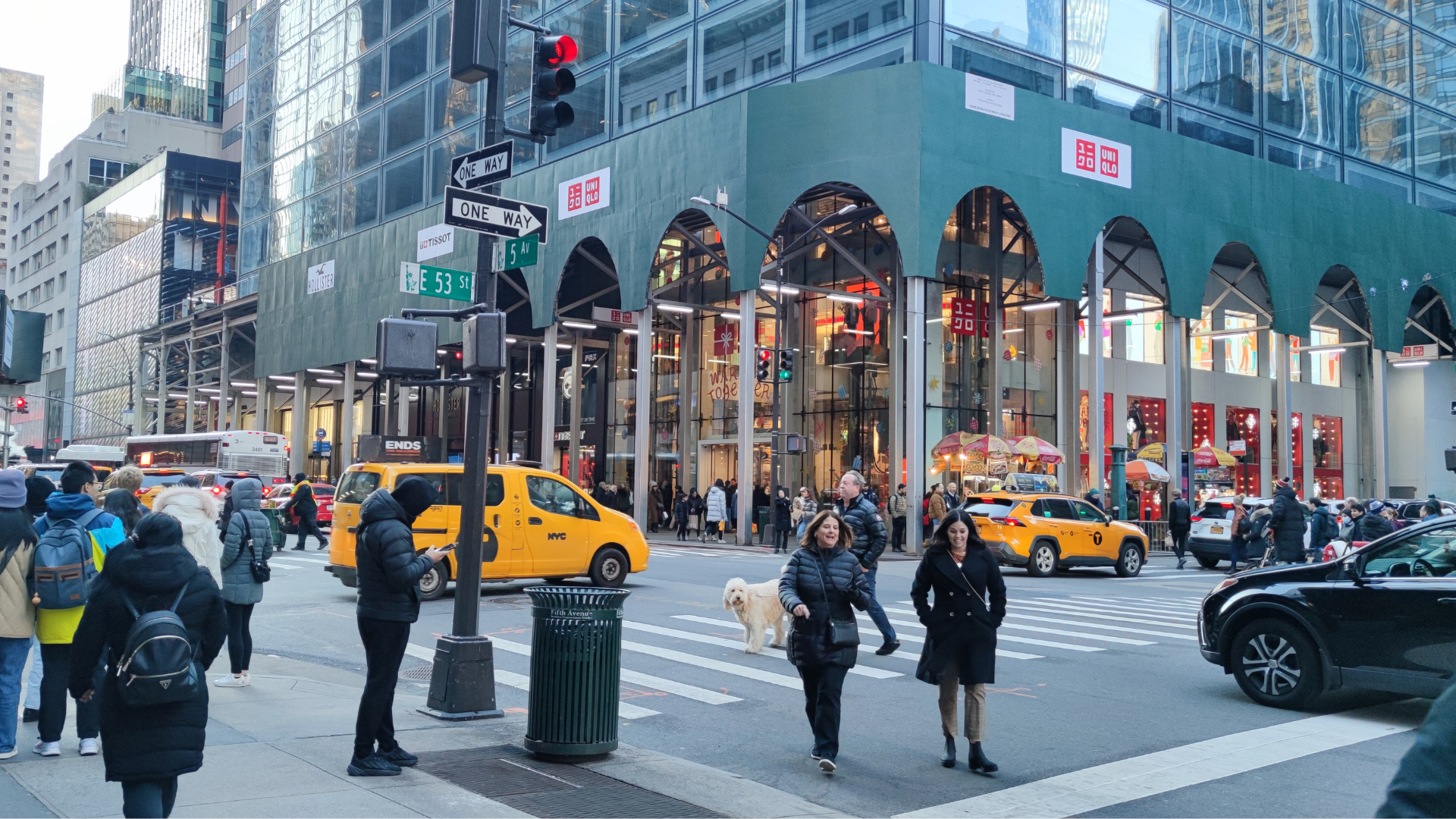 Pedestrians cross a busy intersection in front of a modern glass building featuring green arched storefronts.