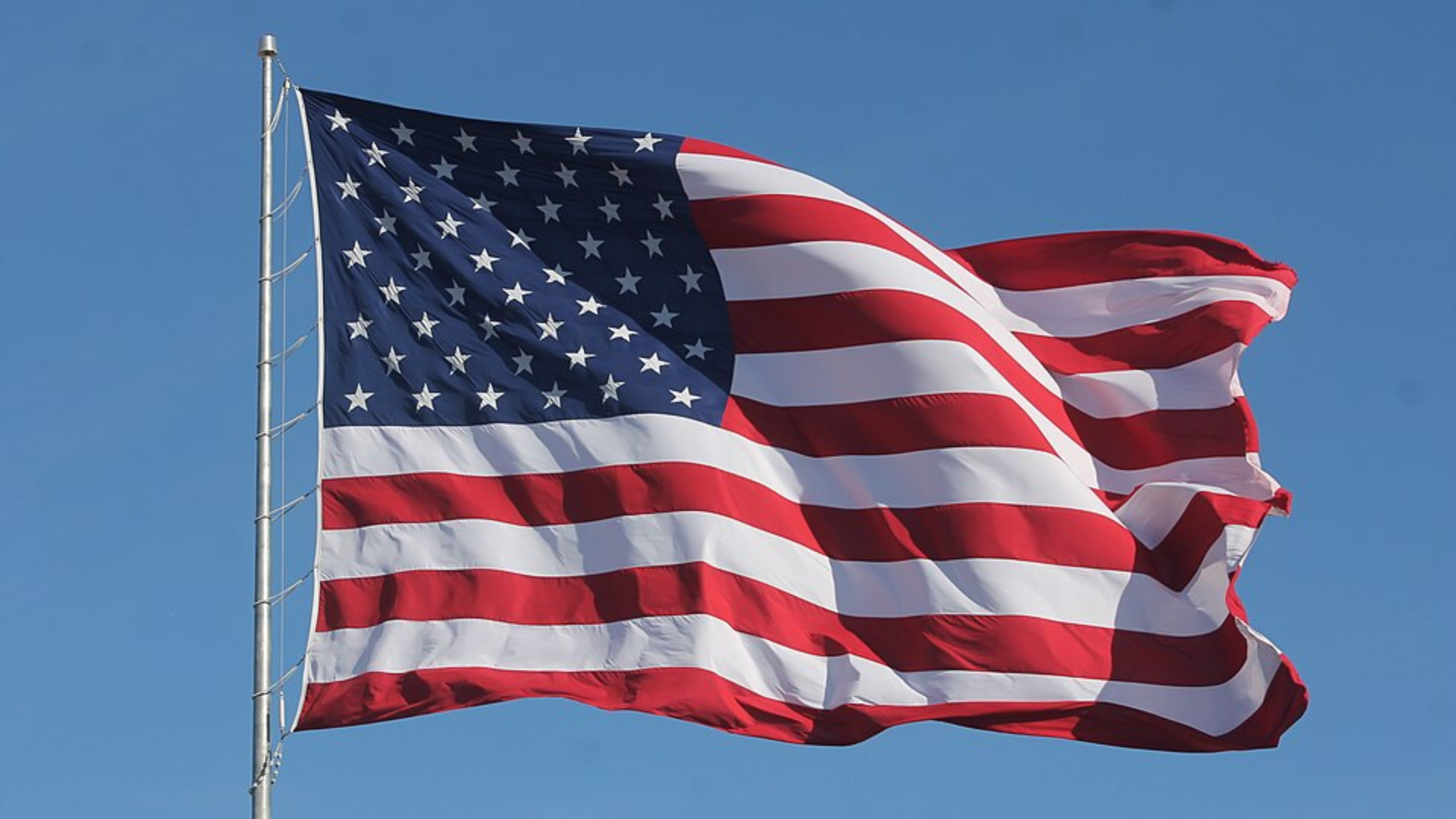 The American flag with stars and stripes waving prominently against a solid blue sky background.