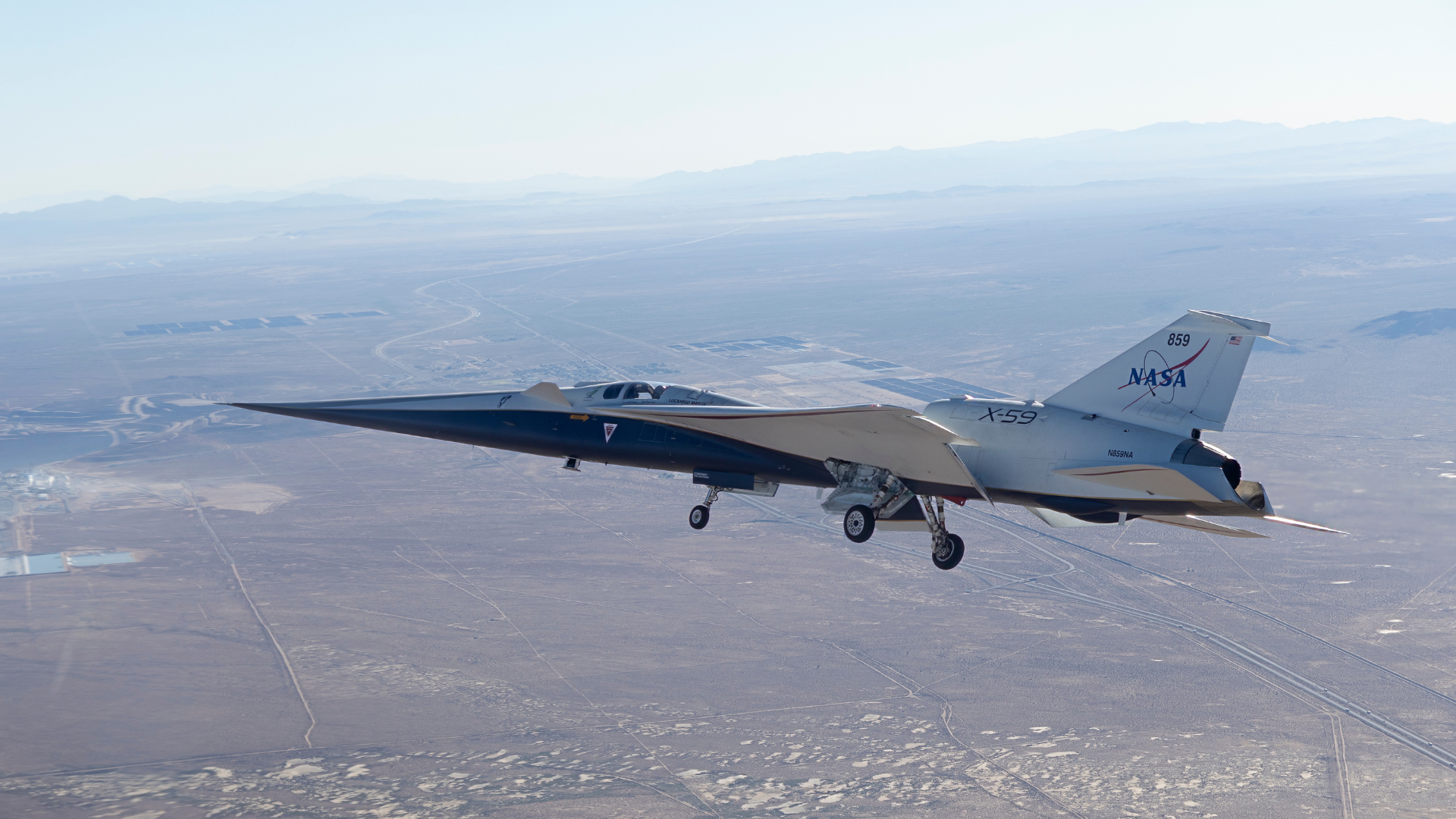 The sleek, long-nosed NASA X-59 experimental aircraft flies with its landing gear down over a vast, arid desert landscape.