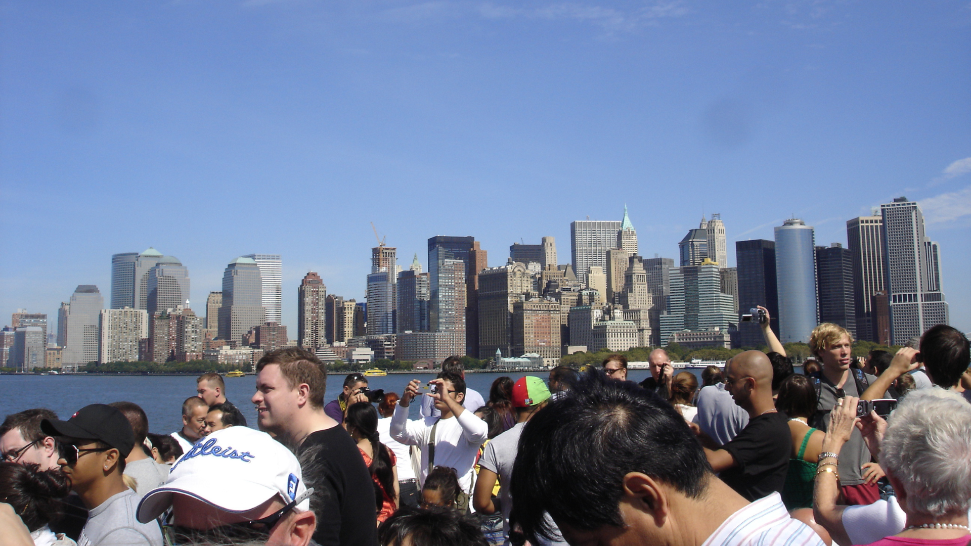 A crowded ferry deck filled with tourists taking photos of the dense Lower Manhattan skyline across the blue water under a clear sky.