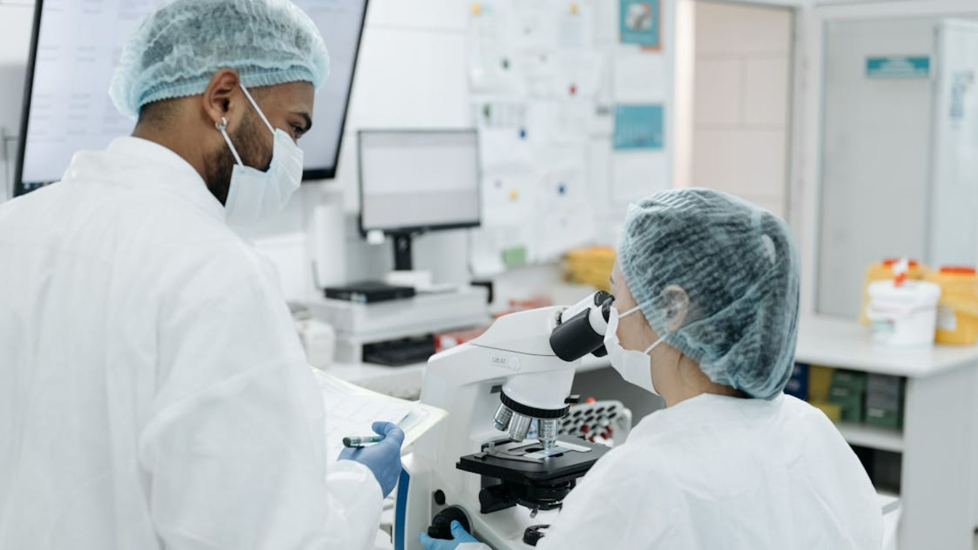 Two scientists in lab coats and hairnets work in a lab.