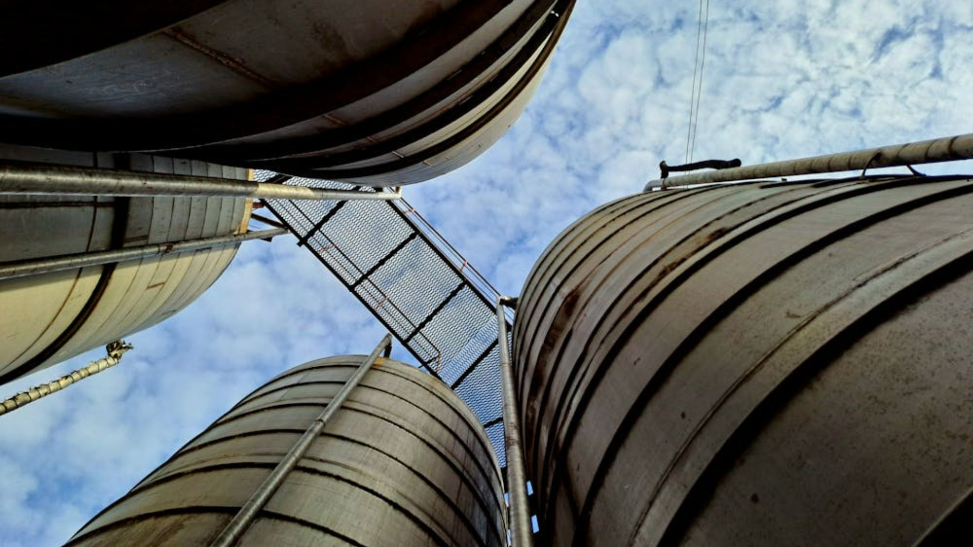 Upward view between large industrial storage tanks and piping against cloudy sky.