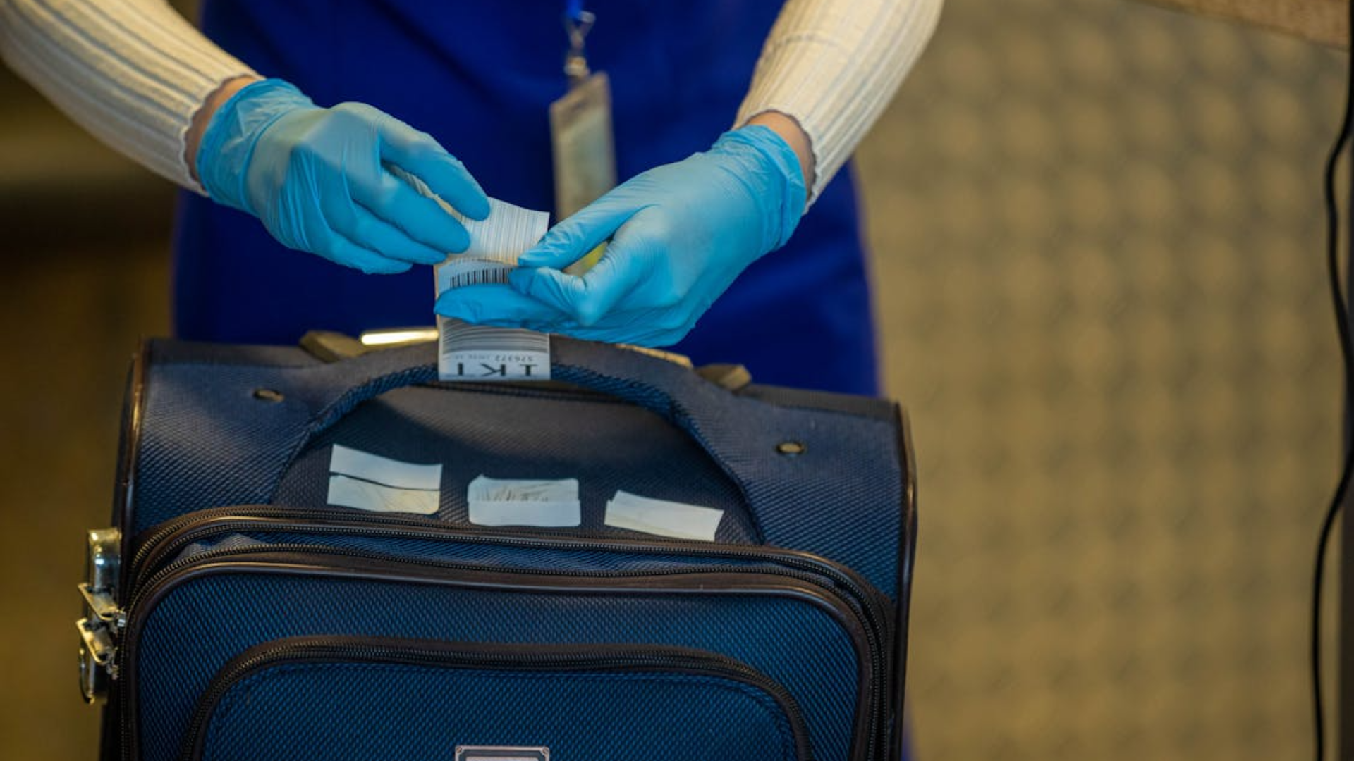 A close-up of an airport staff member in blue gloves attaching a barcode luggage tag to the handle of a blue suitcase.