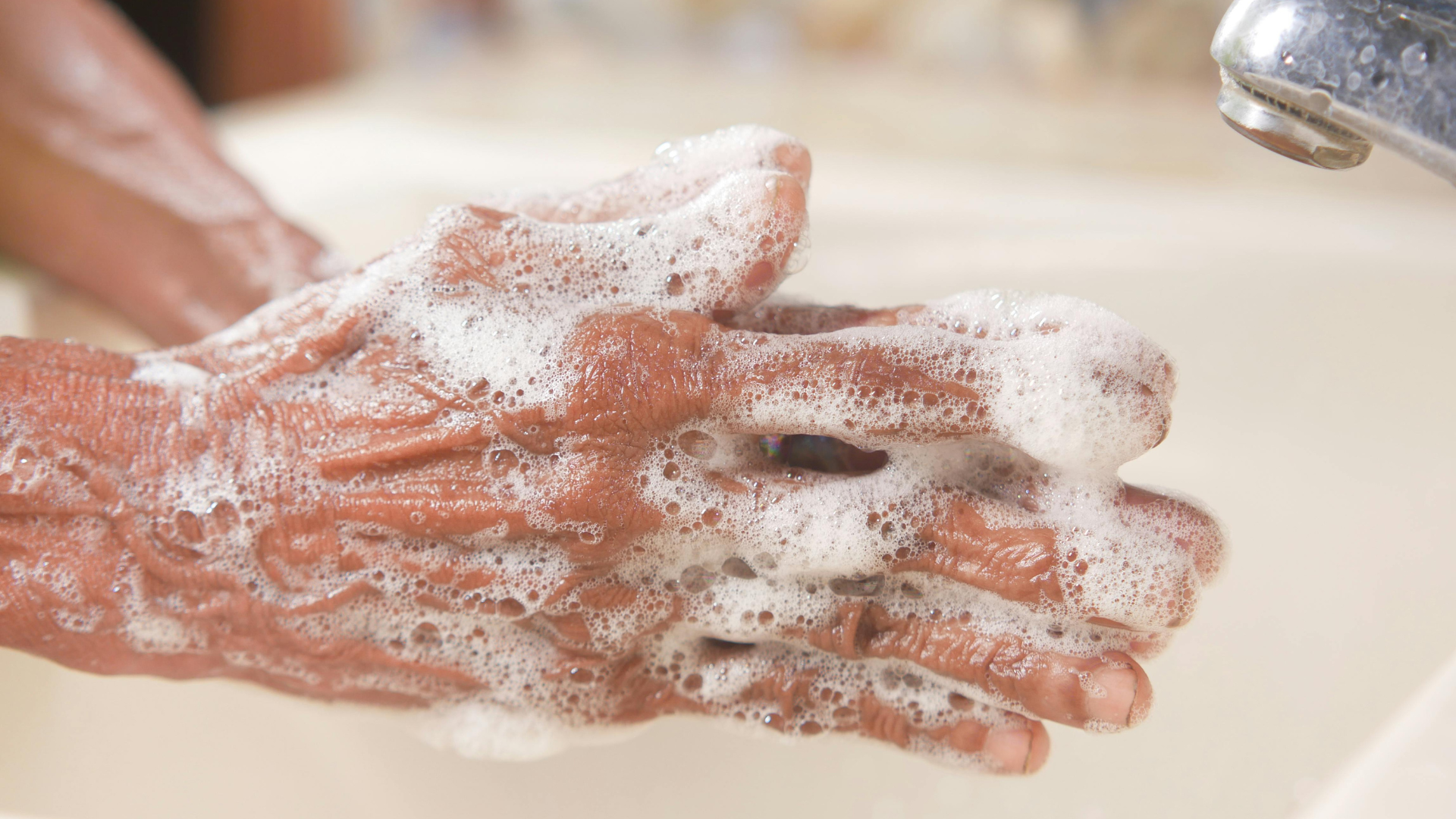 A close-up shot shows hands covered in white soapy bubbles being washed under a chrome faucet.