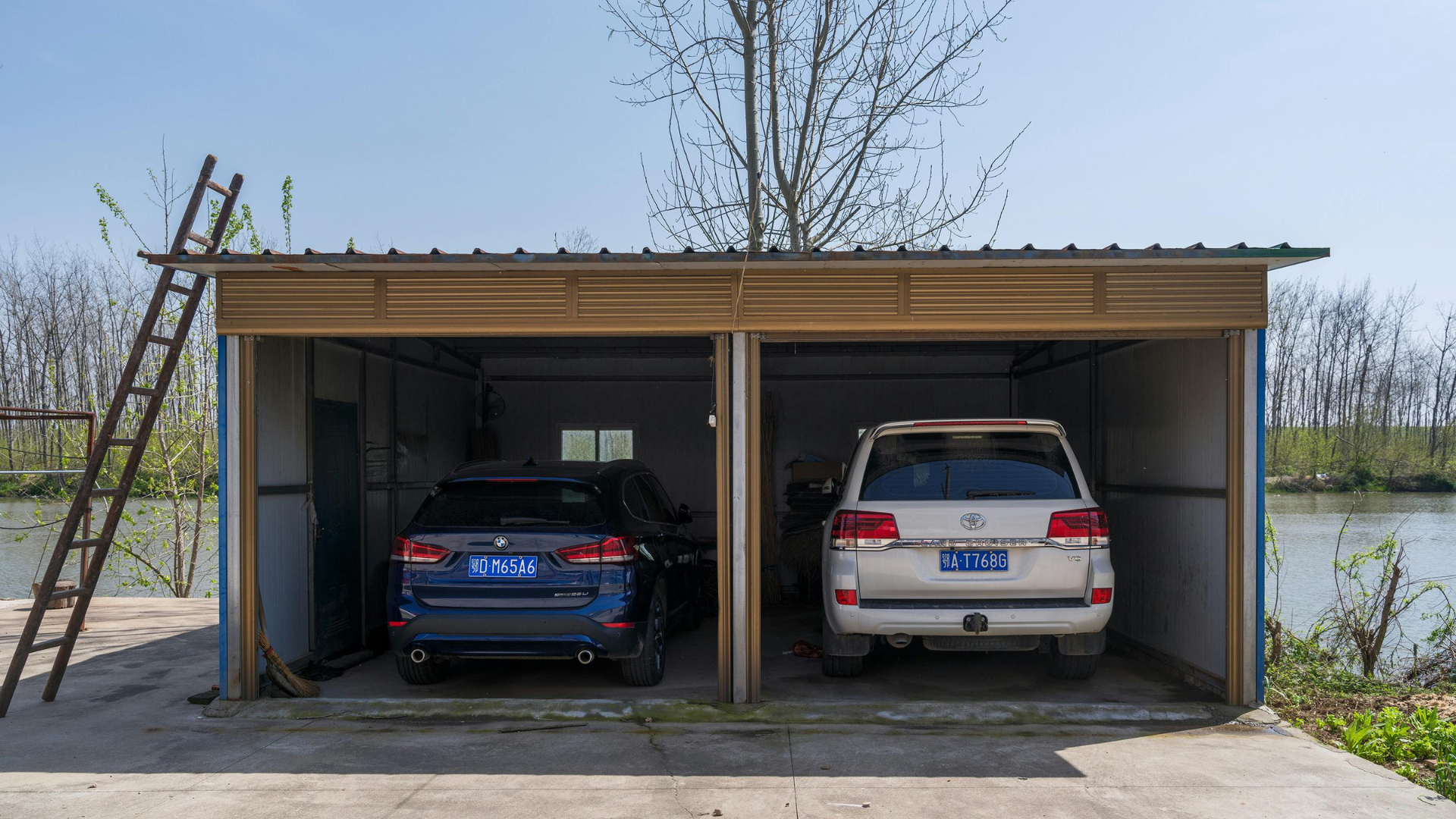 A dark blue SUV and a white SUV are parked side-by-side inside a simple, open-faced corrugated metal garage in a rural outdoor setting.