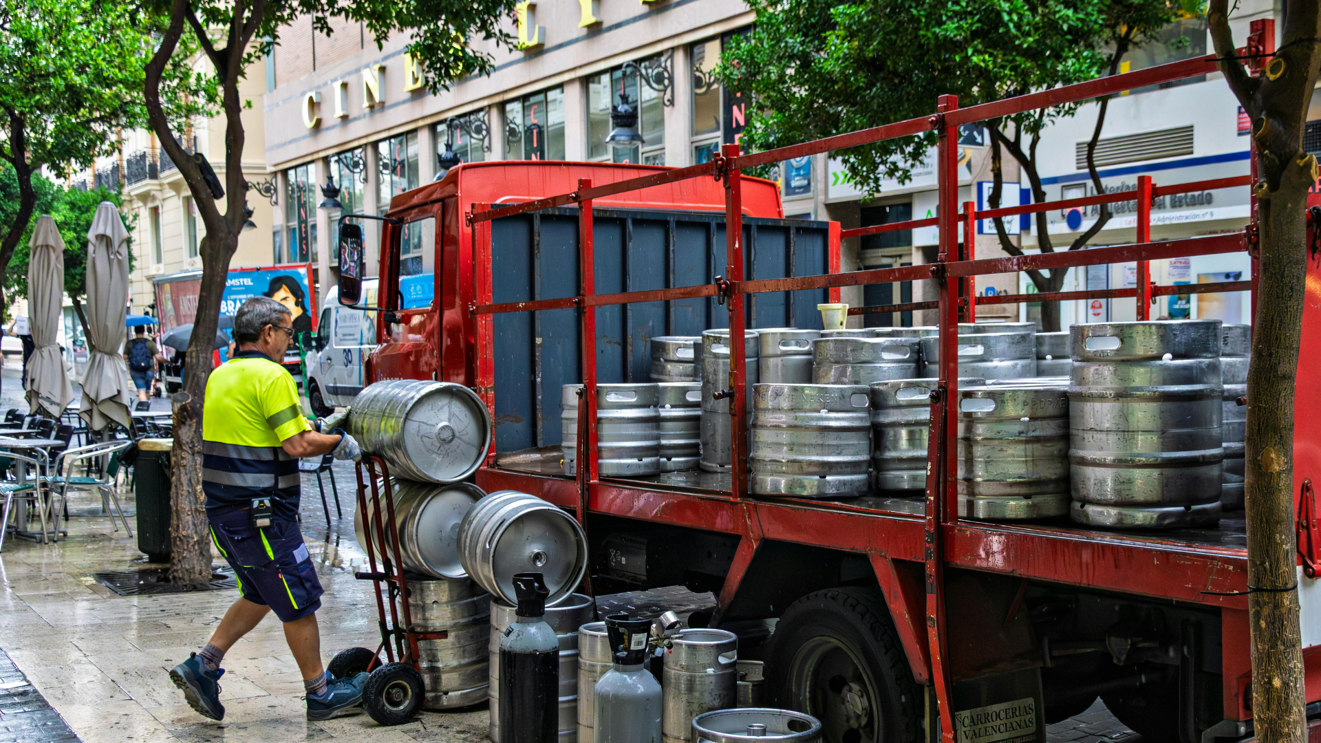  A delivery worker in a high-visibility yellow shirt uses a hand truck to move silver beer kegs next to a red flatbed truck on a city sidewalk.
