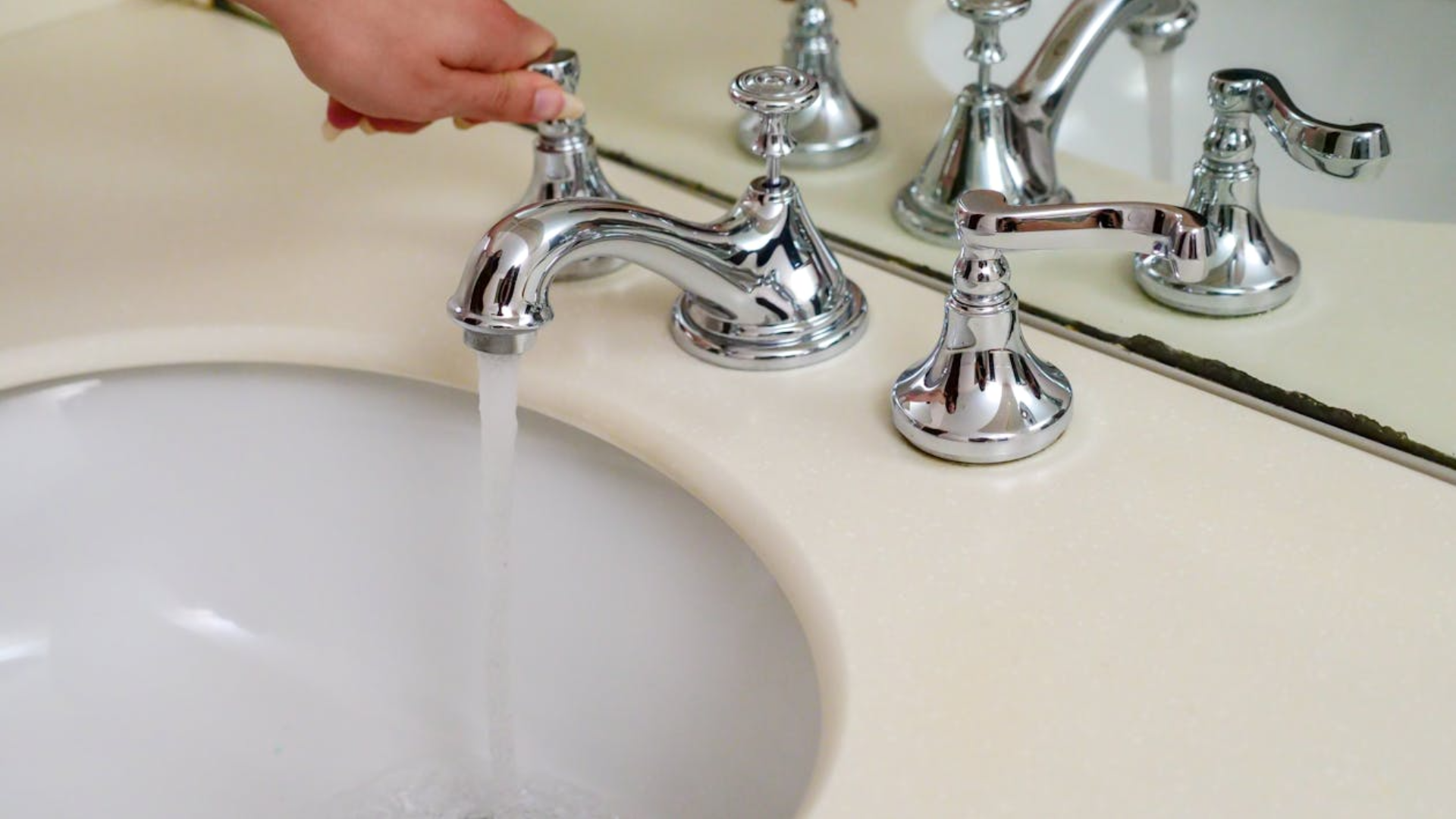  A hand turns the handle of a polished chrome faucet, with water flowing into a white bathroom sink.