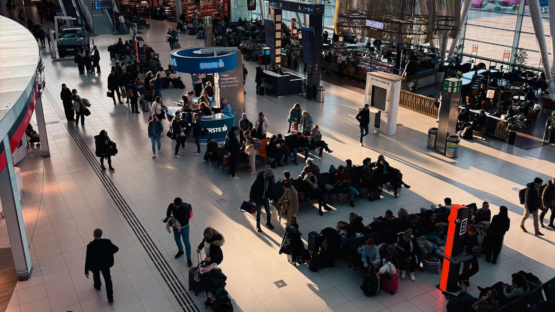 A high-angle shot captures the bustling interior of a modern airport terminal with travelers walking through a spacious, sunlit hall filled with seating areas and shops.