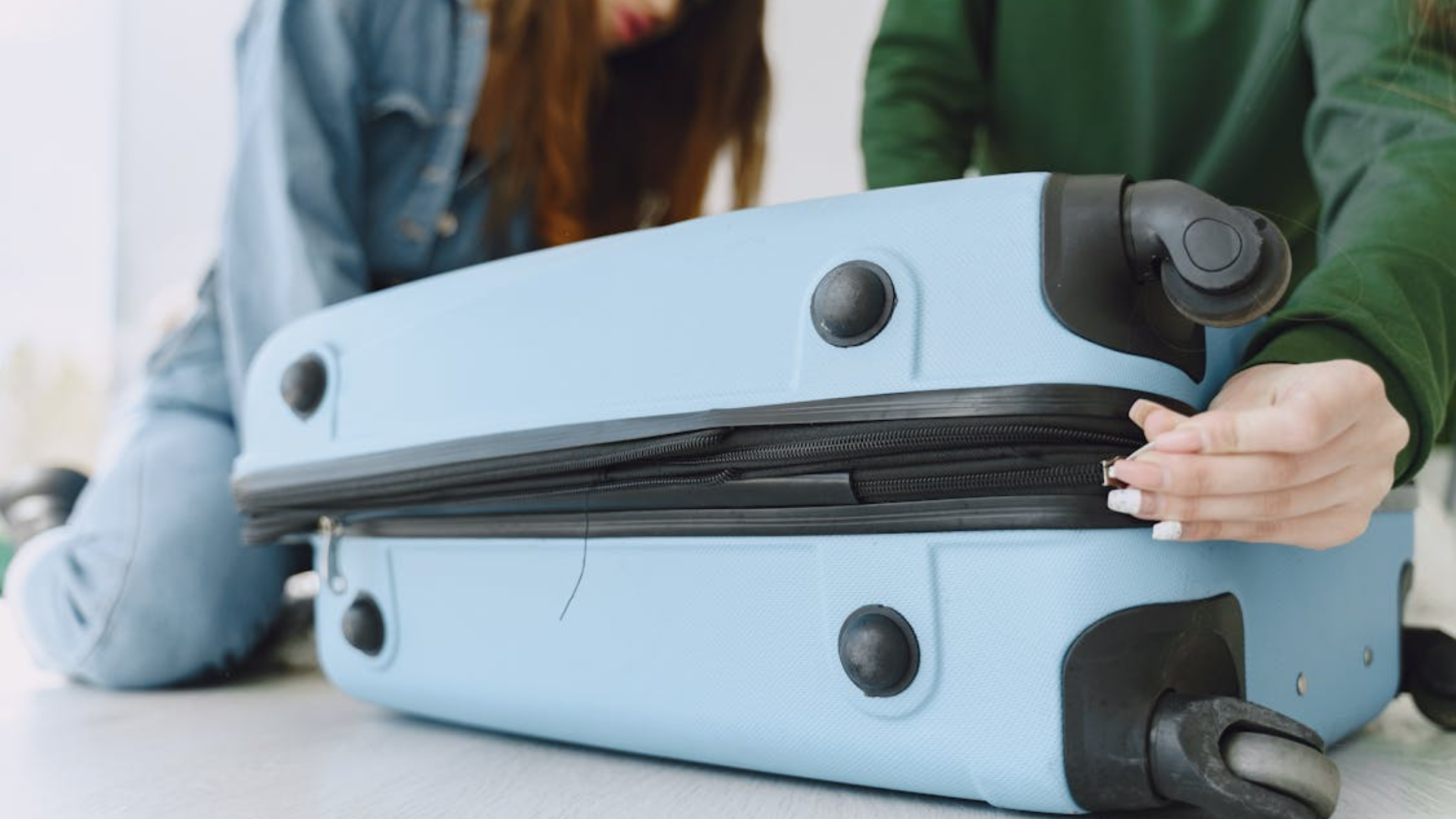 A low-angle shot of a person zipping up a light blue hardshell suitcase on the floor.