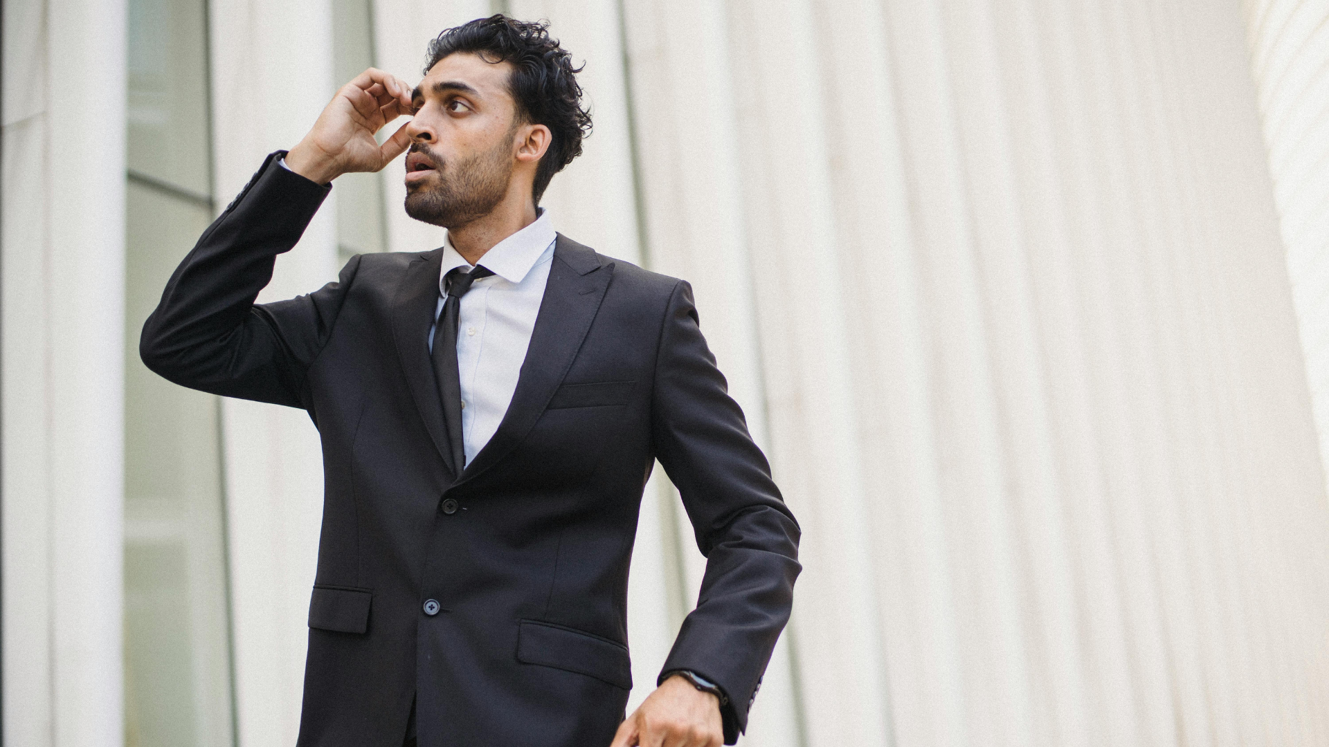 A man in a black suit and tie looks off-camera with a concerned expression while in front of a modern white building.