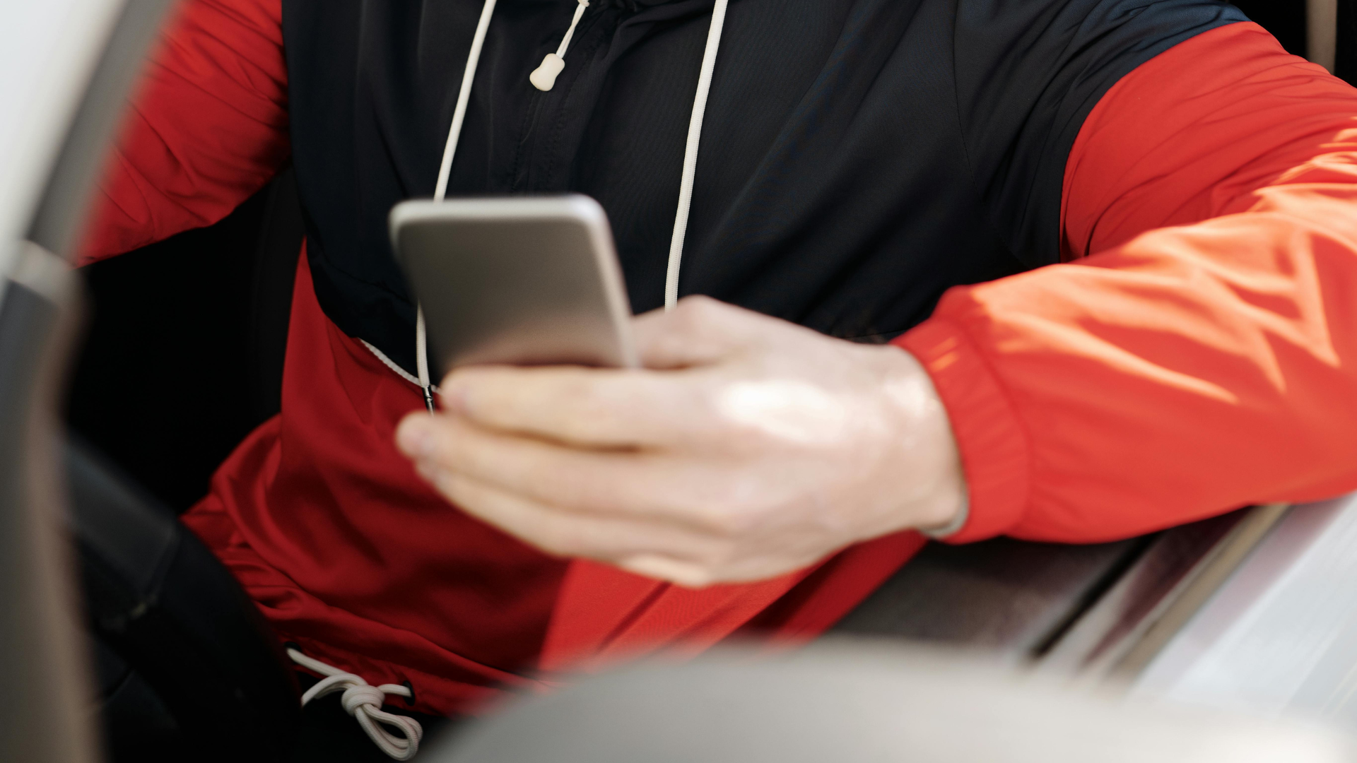 A man wearing a red and black jacket sits in the driver's seat of a car while looking down at his smartphone.