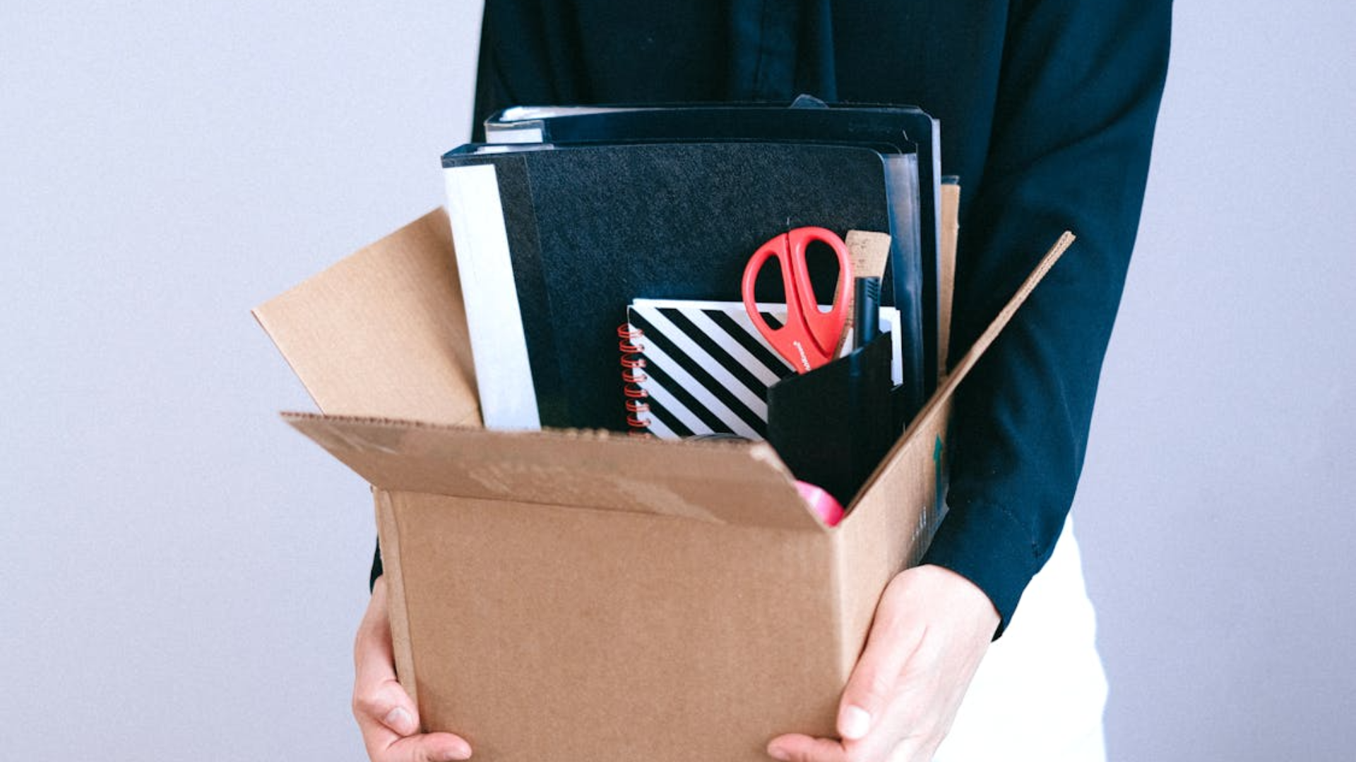  A person in dark clothing holding a cardboard box filled with office supplies, including folders, notebooks, and scissors.