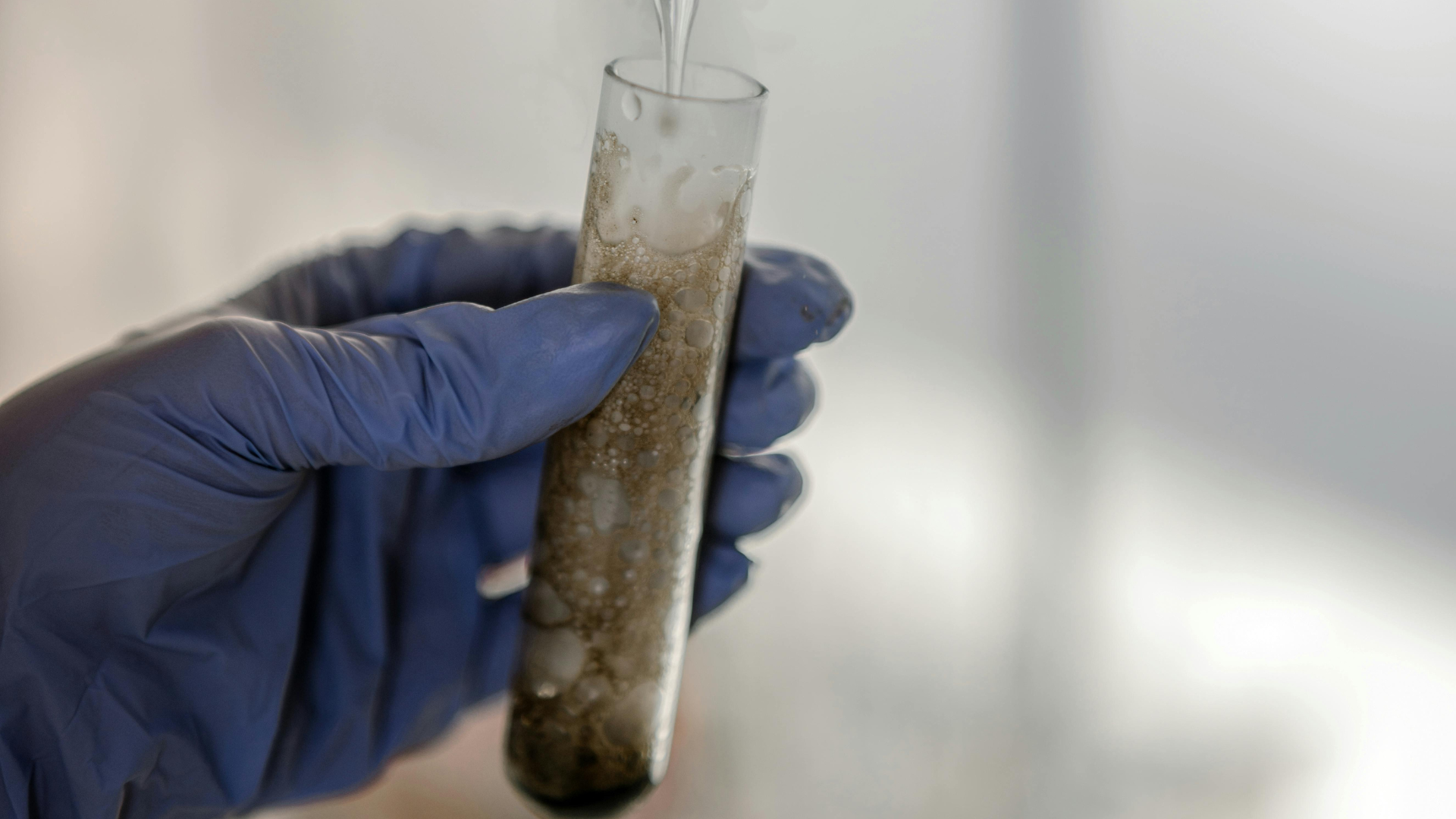 A person wearing blue gloved hands holds a test tube filled with a bubbling brown liquid that is emitting white vapor.