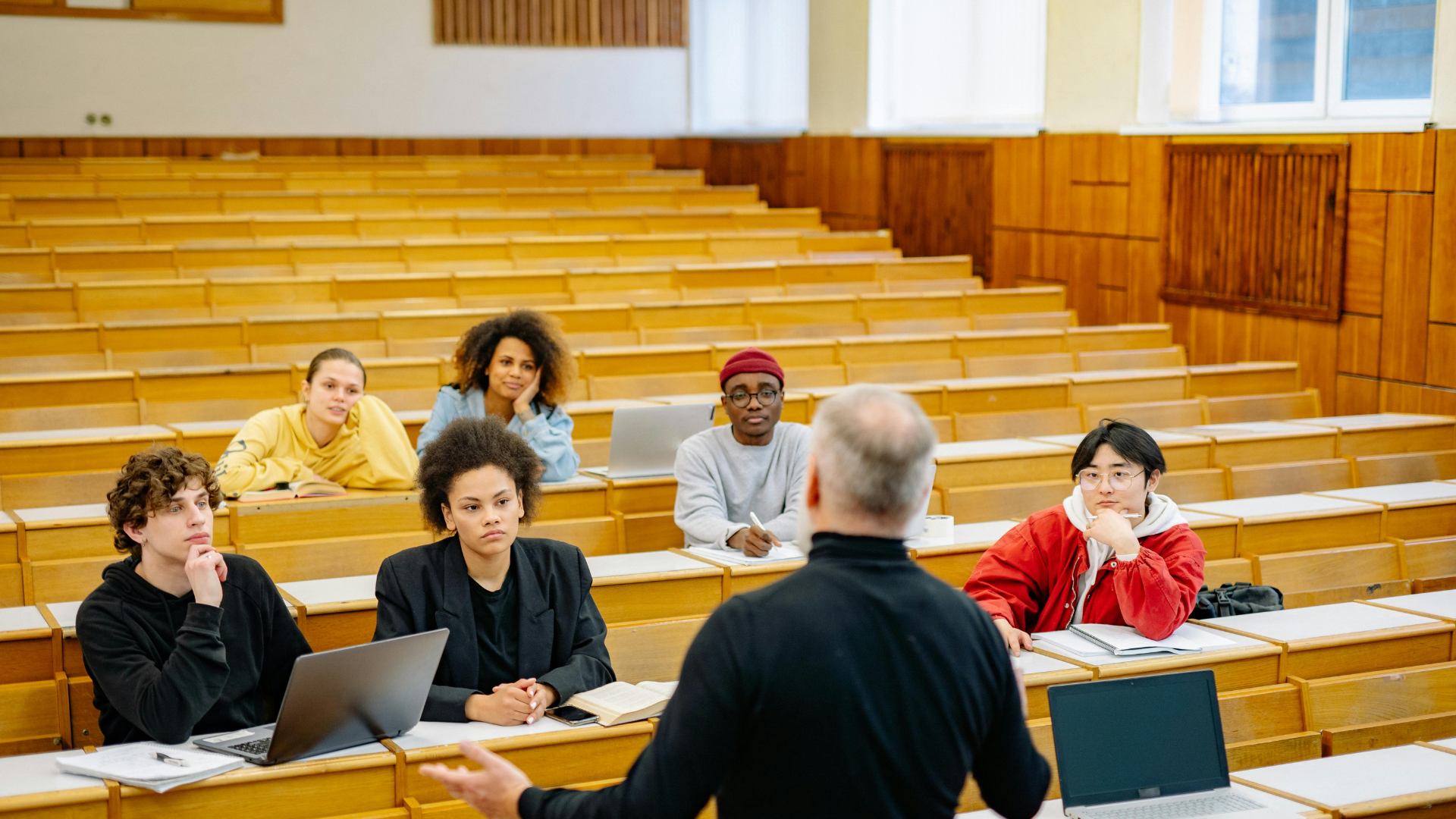 A professor stands at the front of a large, wood-paneled lecture hall addressing a small group of attentive students.