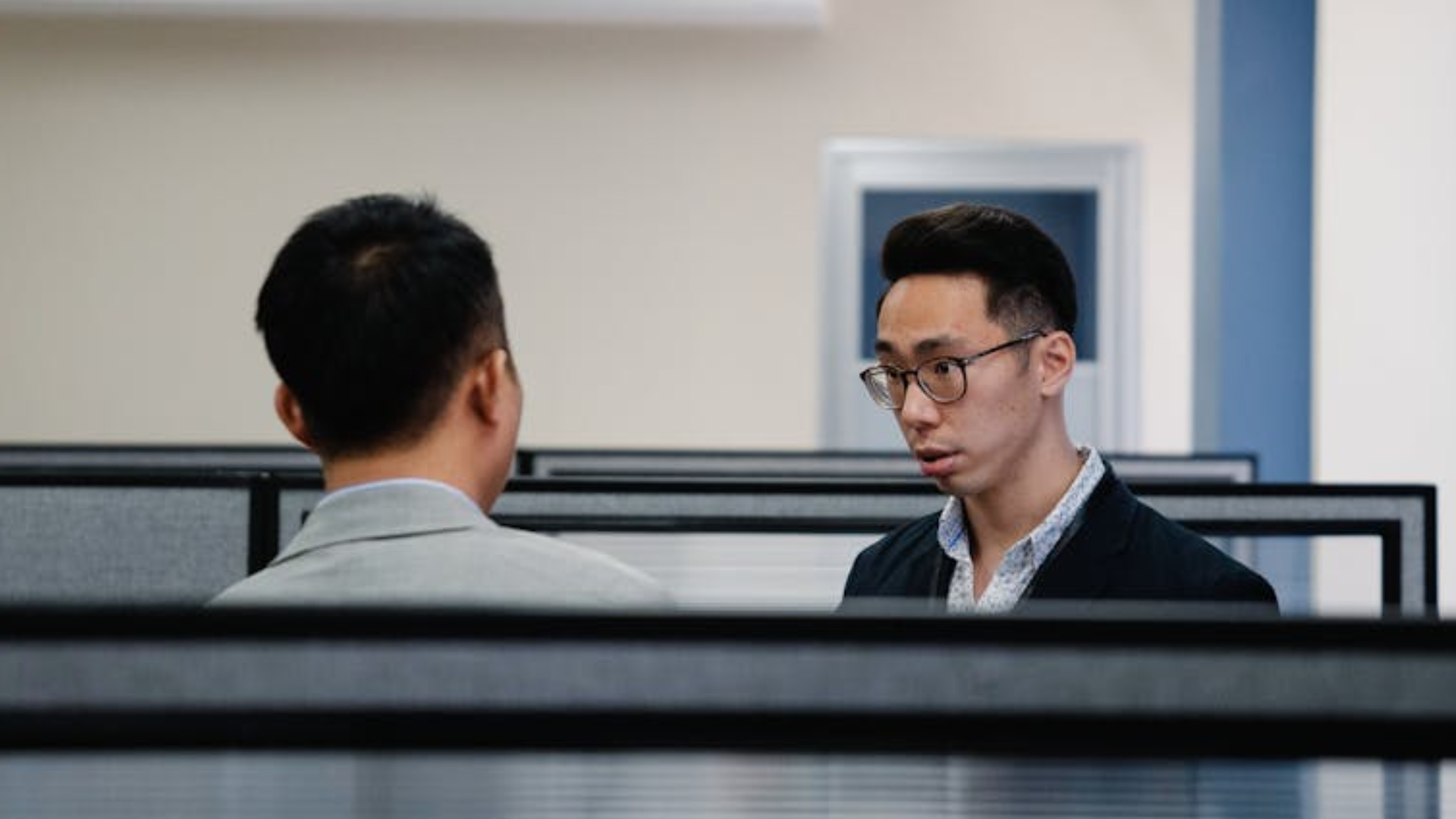  A profile shot of two men in a modern office, one wearing glasses and looking attentively at the other in a cubicle setting.