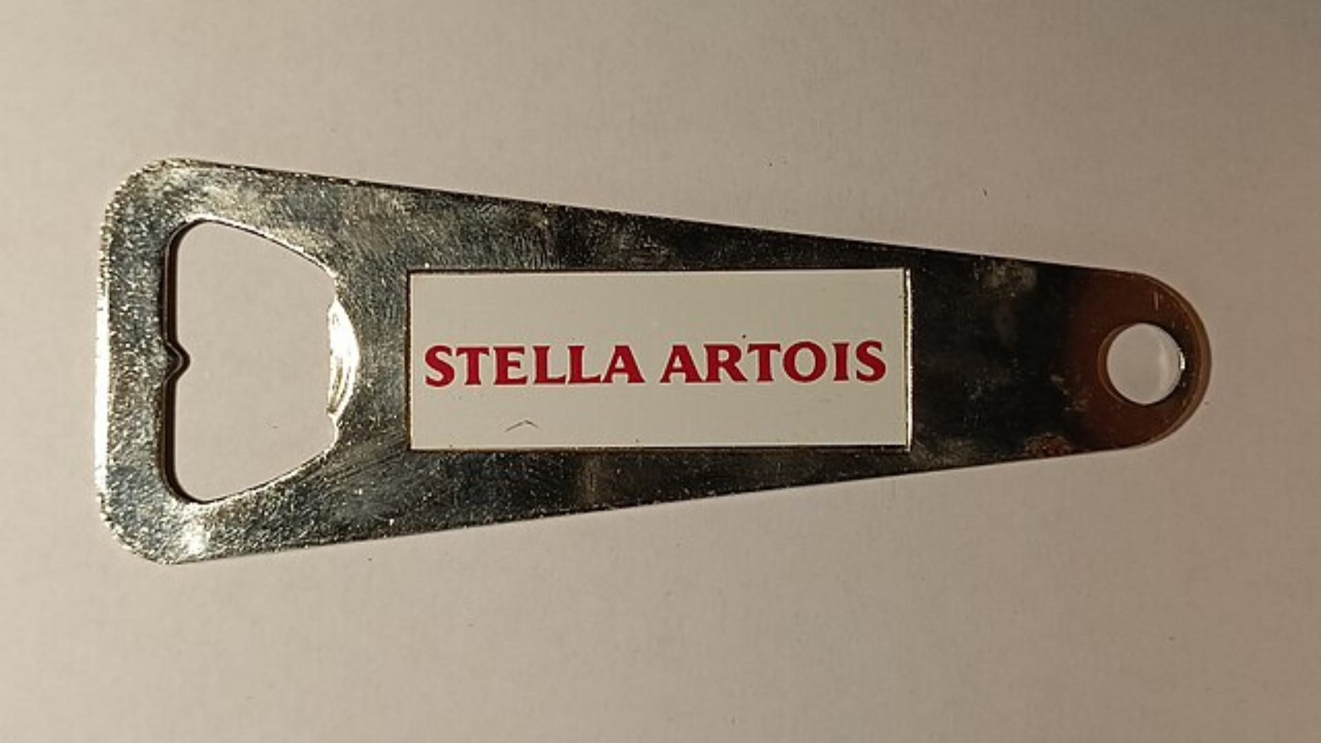 A top-down shot of a metallic, flat-handled bottle opener featuring the red Stella Artois logo on a white rectangular background.