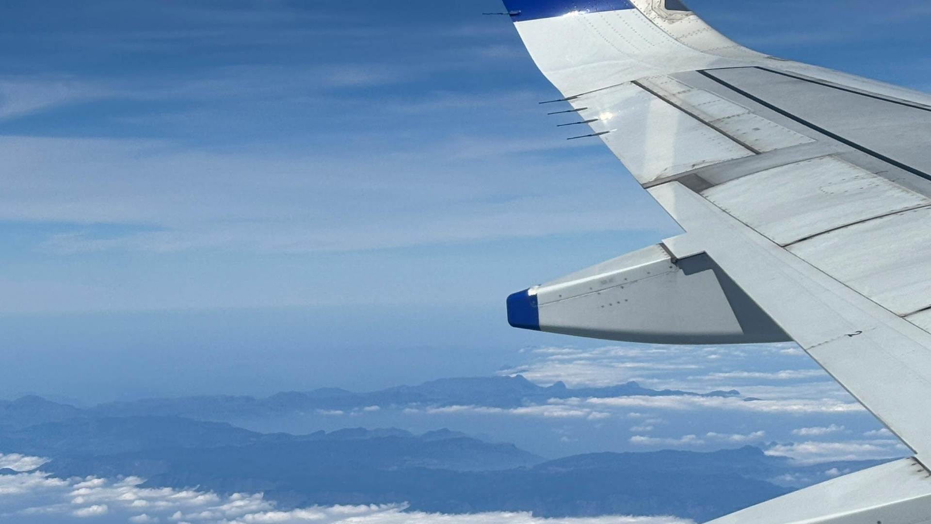 A view from an airplane window shows a white wing with blue tips soaring above a vast layer of fluffy white clouds under a clear blue sky.