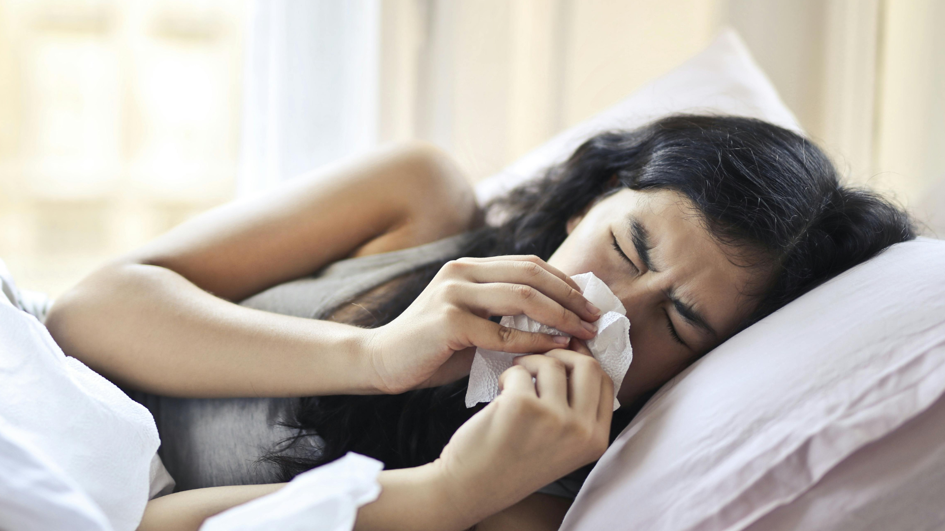A woman lies in bed under a white duvet, using a tissue to blow her nose while appearing unwell.