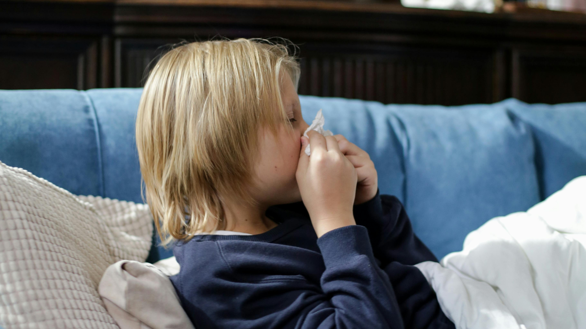 A young child with blonde hair sits on a blue sofa, wrapped in a white blanket and blowing their nose with a tissue.
