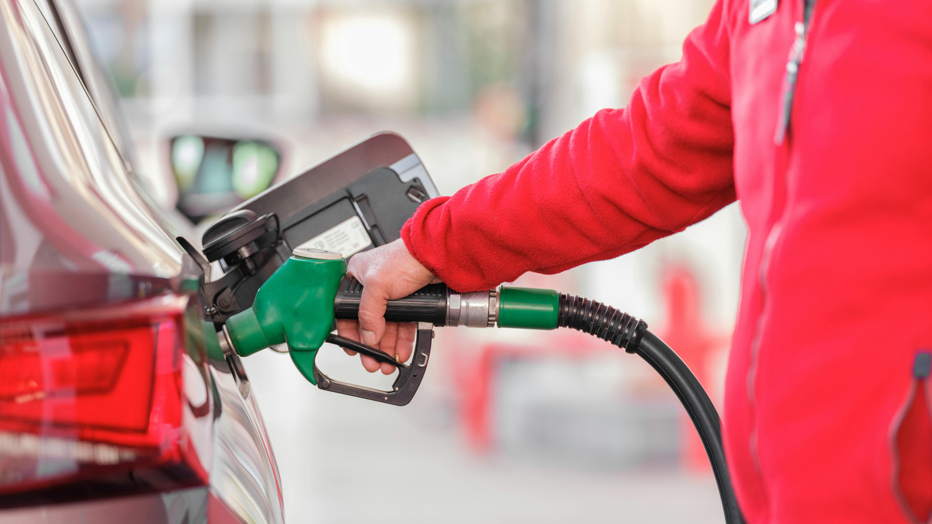 An attendant in a red jacket inserts a green fuel nozzle into the gas tank of a grey car.