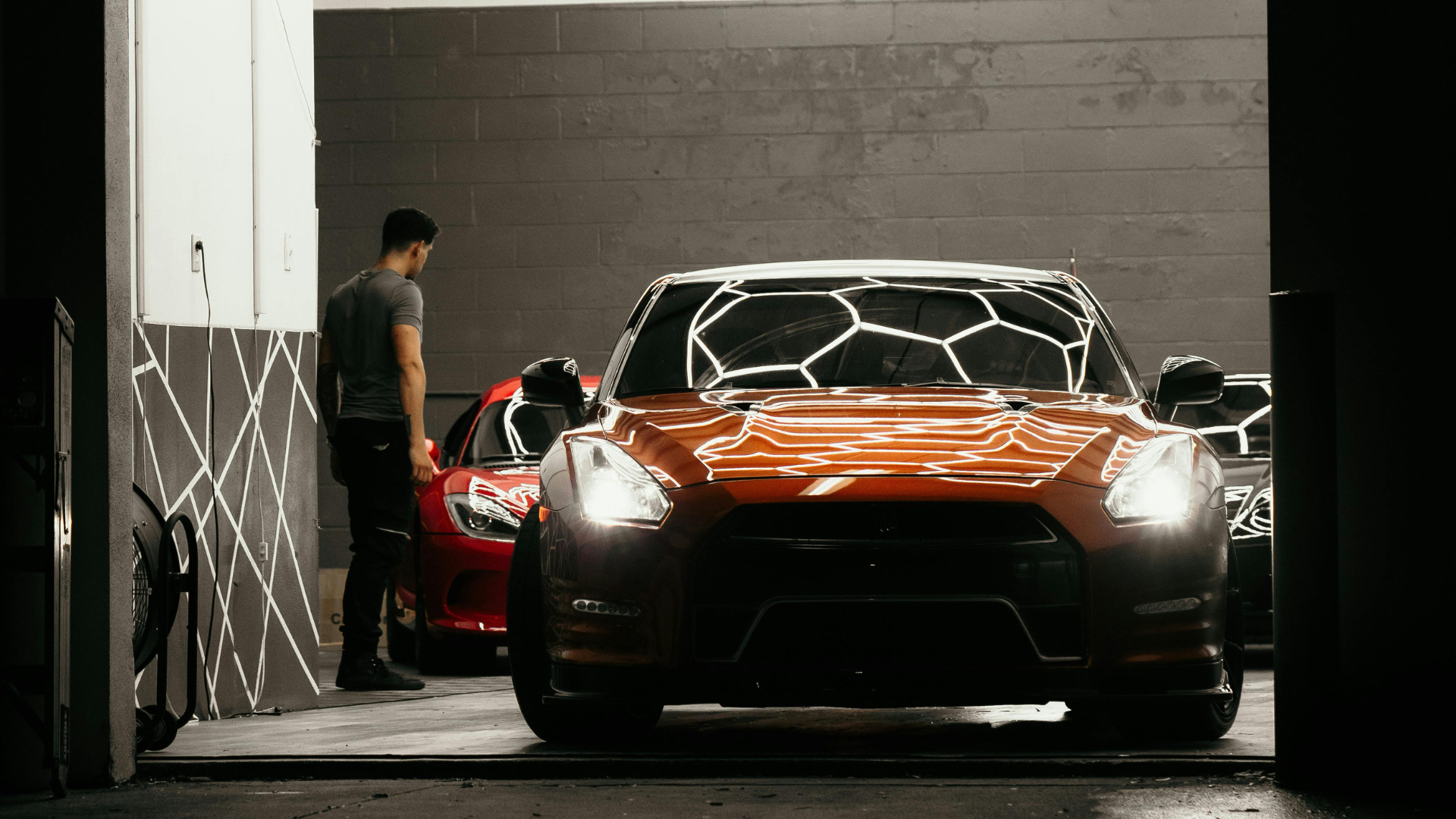 An orange sports car with glowing headlights is parked inside a well-lit garage, with a person standing in the background near a red vehicle.
