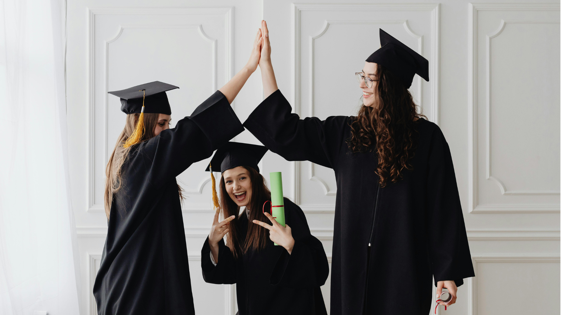 Three graduates in black caps and gowns celebrate their achievement by high-fiving and holding their diplomas.