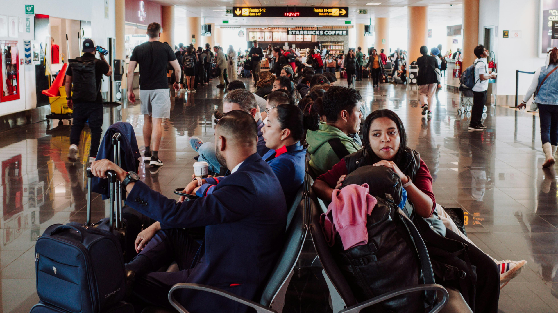 Travelers wait in rows of silver seats within a brightly lit airport terminal, with some standing near a Starbucks in the background.