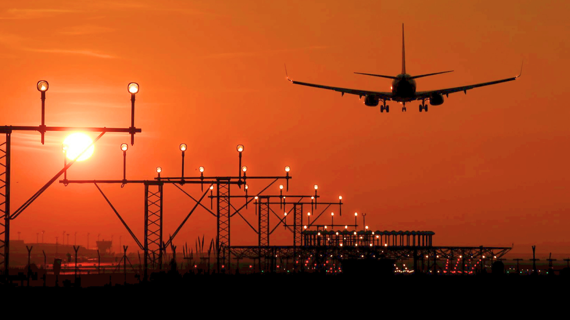 An airplane is captured in silhouette as it descends toward a runway lined with glowing approach lights against a deep orange sunset..