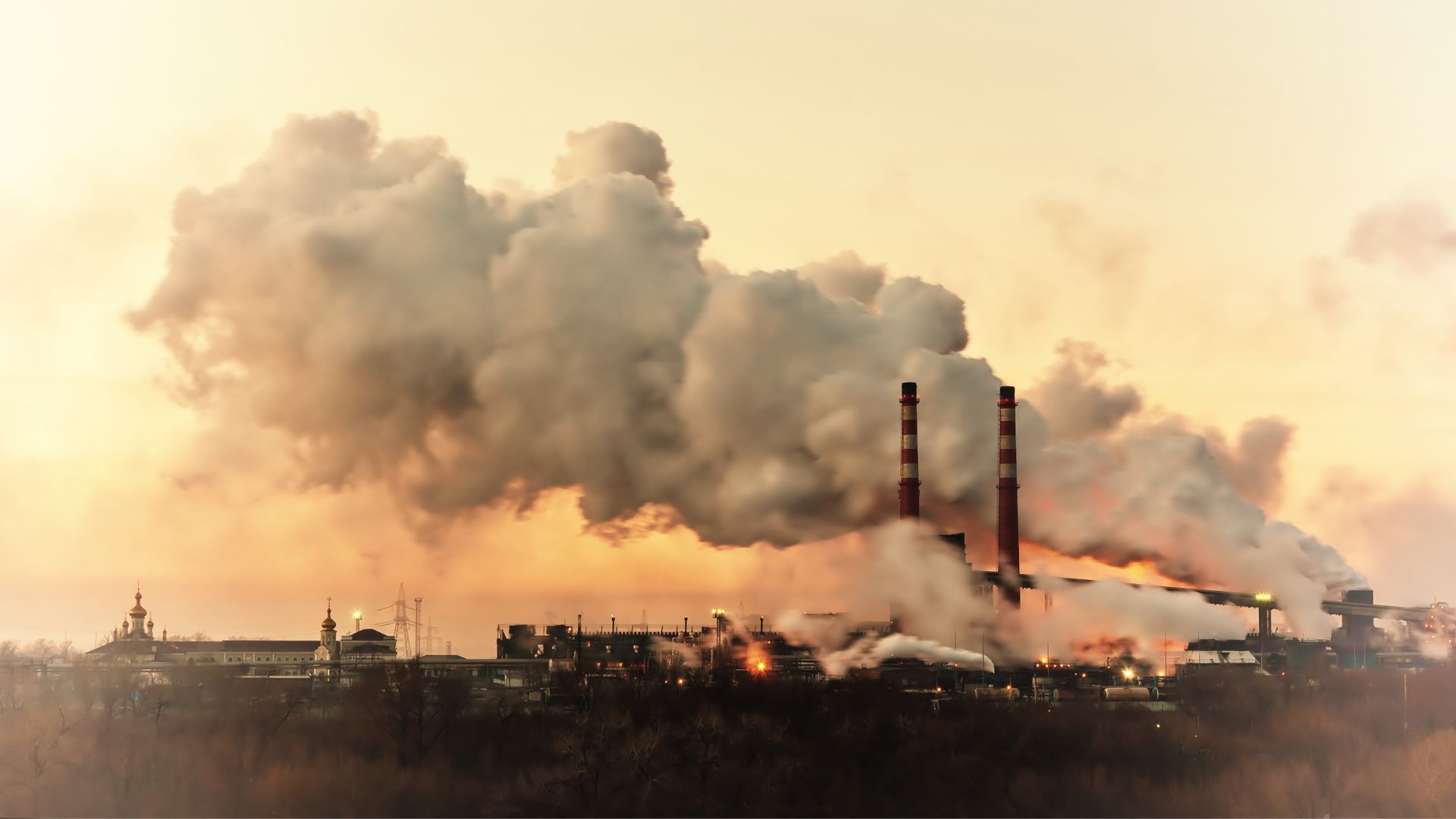 Industrial landscape with two tall smokestacks emitting thick plumes of smoke into the sky at sunset.