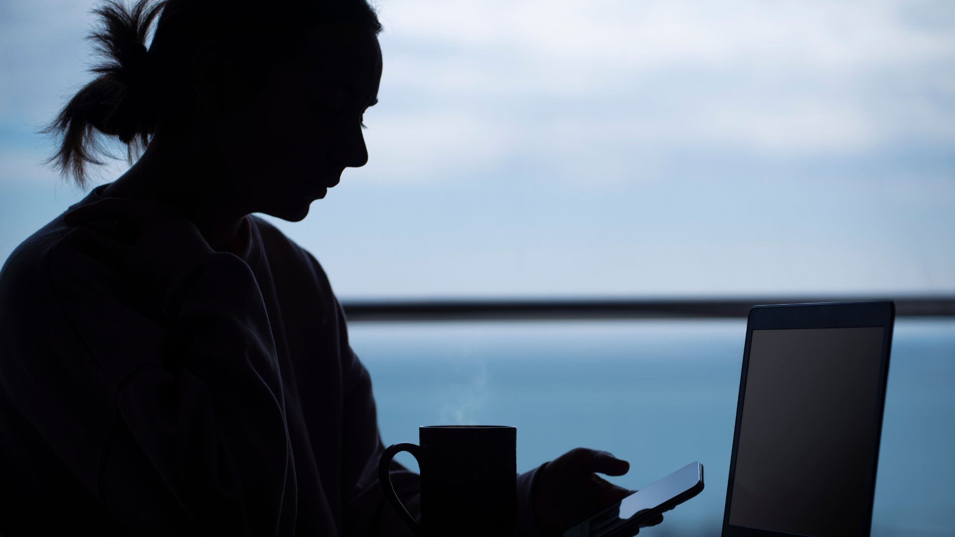 Silhouetted woman at a table using a smartphone with a laptop nearby.