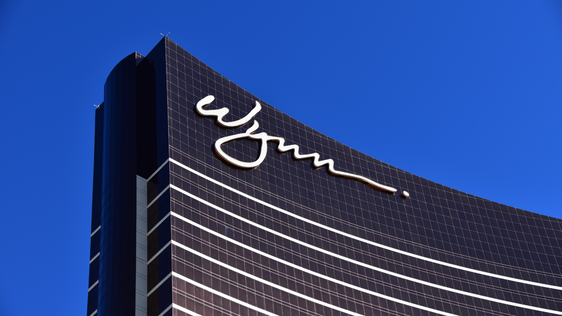 A low-angle close-up of the top of the Wynn Las Vegas hotel, featuring its curved bronze glass facade and white signature logo against a bright blue sky.
