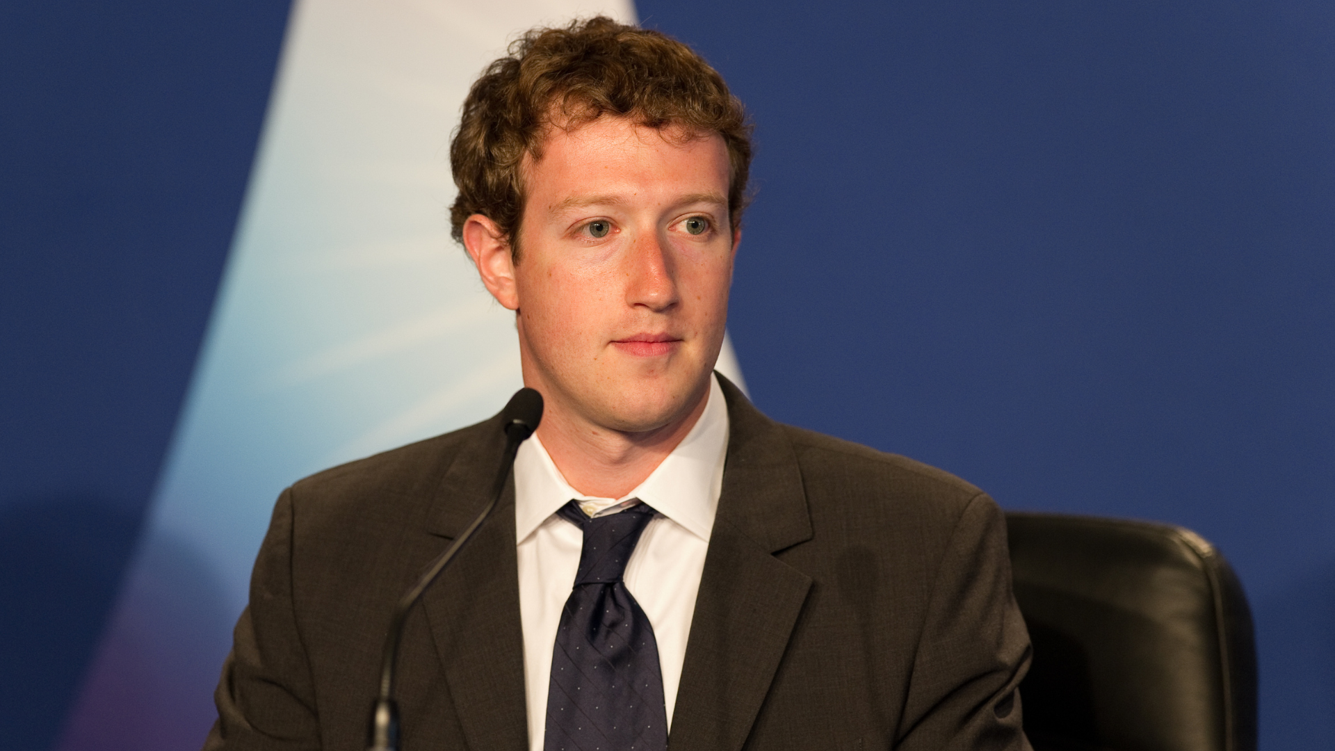 A professional portrait of Mark Zuckerberg in a dark suit and blue tie, looking attentively toward a speaker.