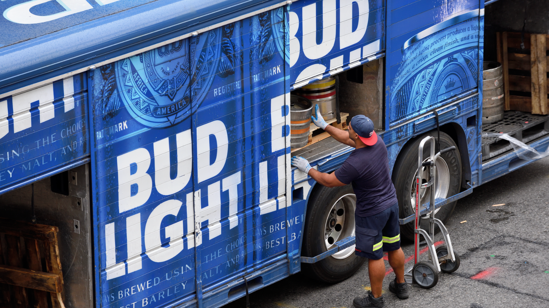 An overhead view of a delivery worker in a blue shirt and red cap unloading metal kegs from a blue Bud Light delivery truck.