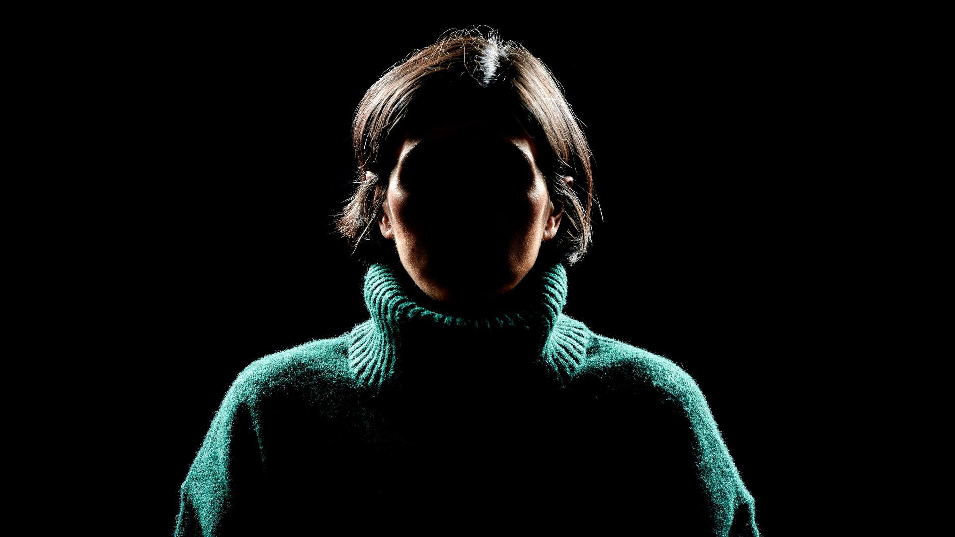 A person in a green sweater faces forward in low lighting, casting their face into shadow against a black background.