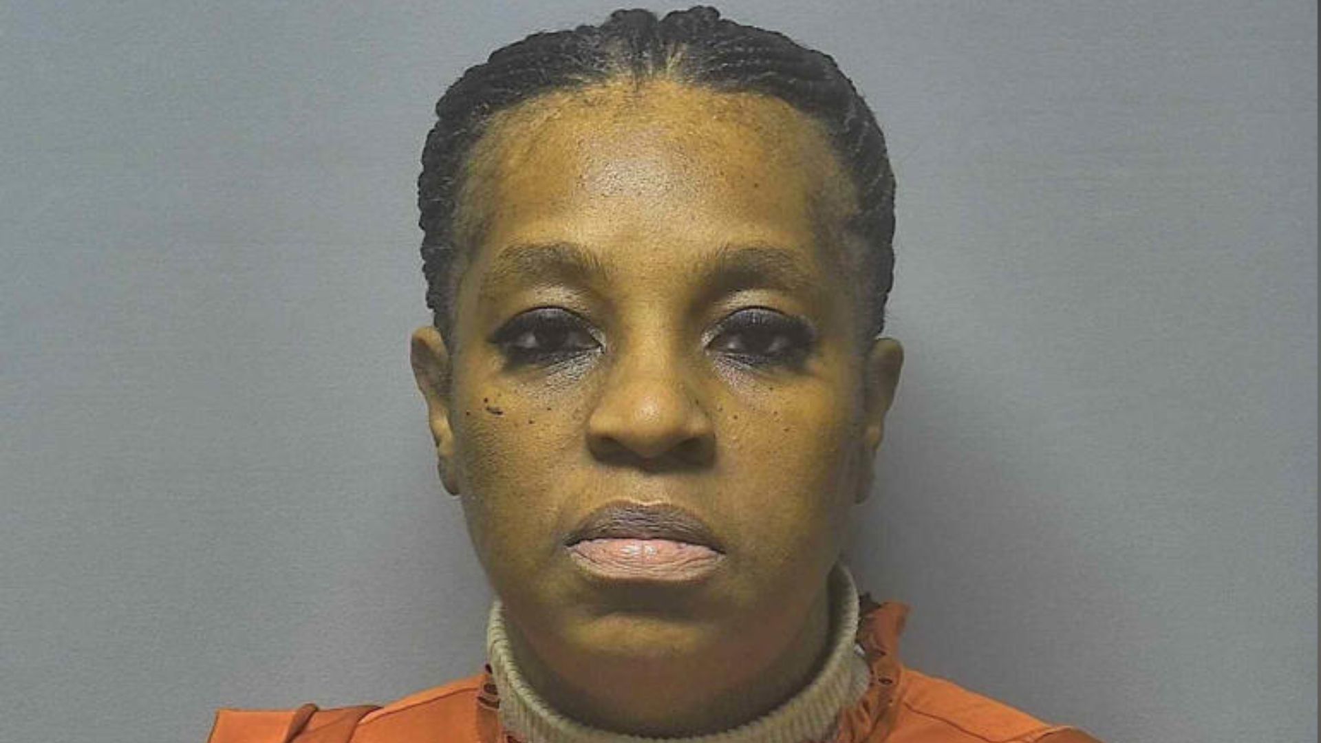 Woman in orange jumpsuit mugshot facing camera against neutral background.