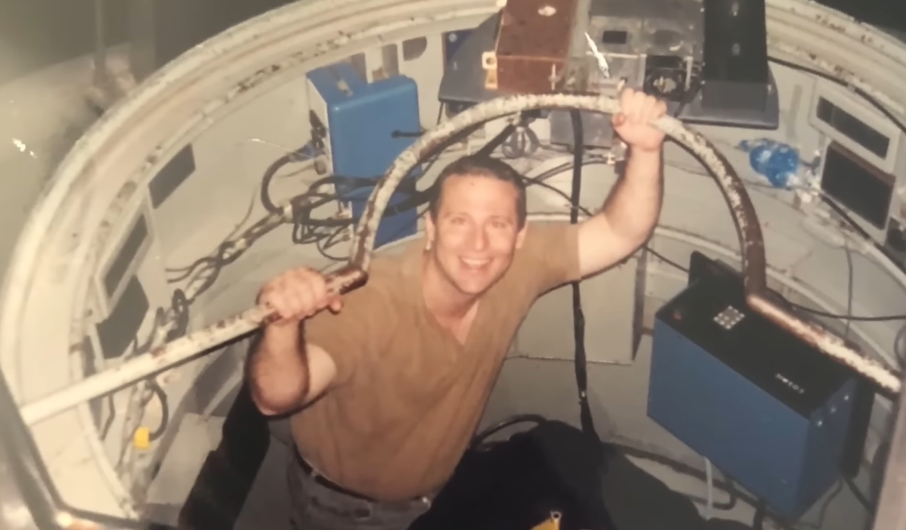Michael David Hicks smiling while holding metal bar inside confined industrial or experimental chamber.