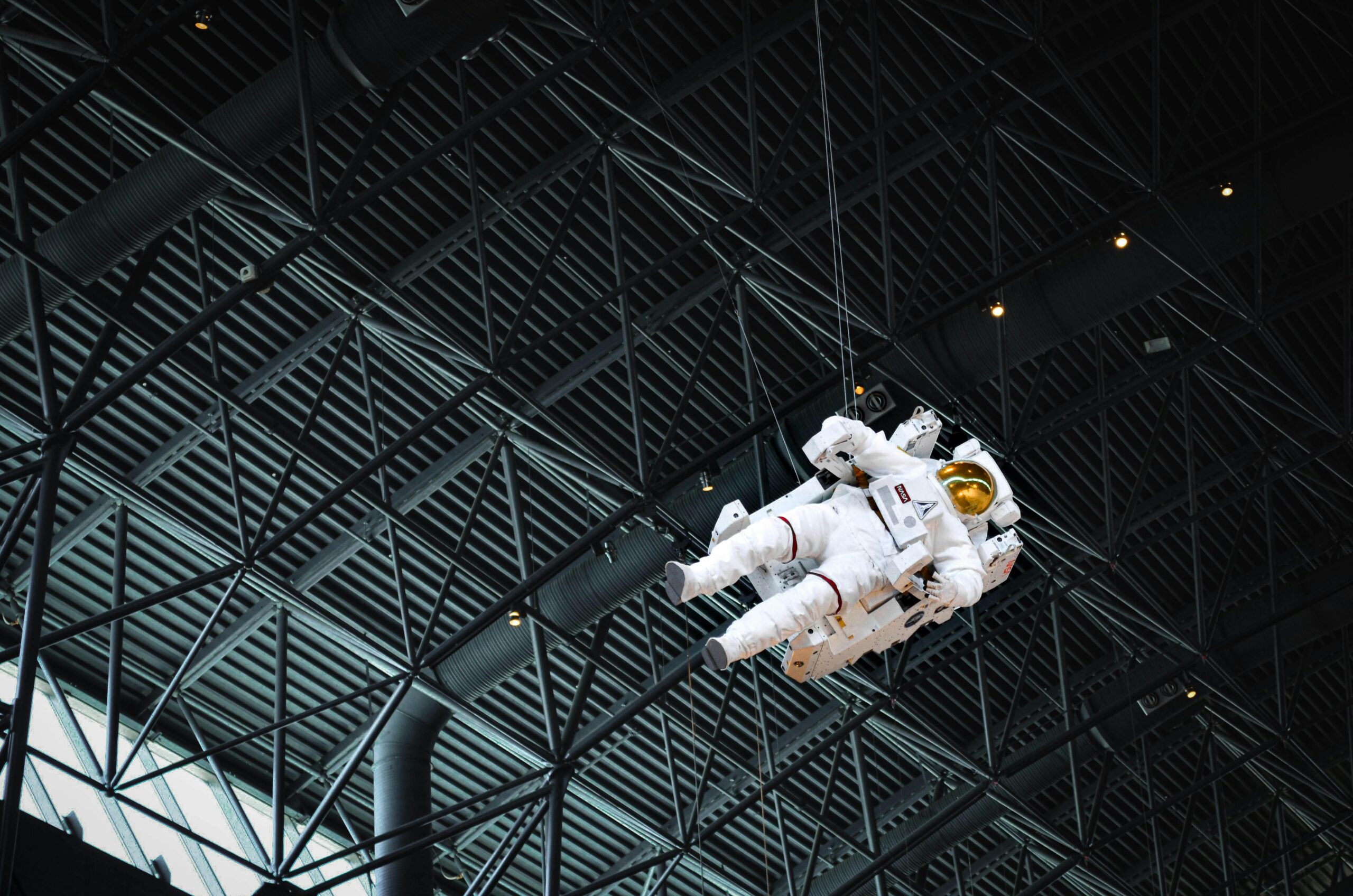 Astronaut model suspended from ceiling inside building with metal framework overhead.