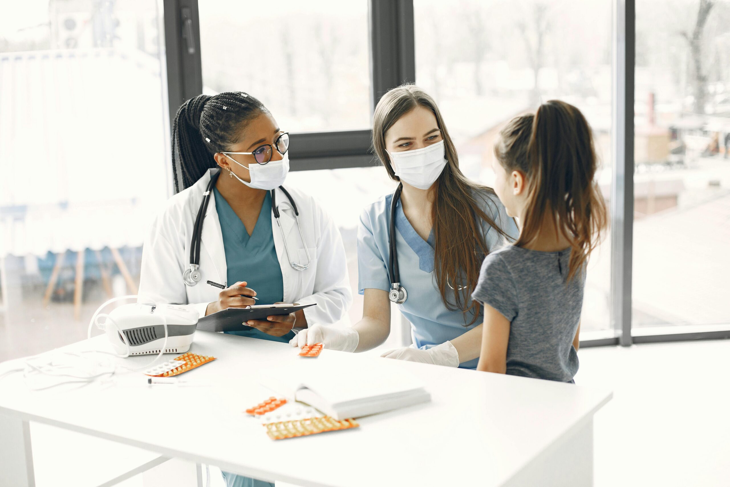 Two healthcare workers wearing masks speak with a young girl while reviewing notes at a clinic table.