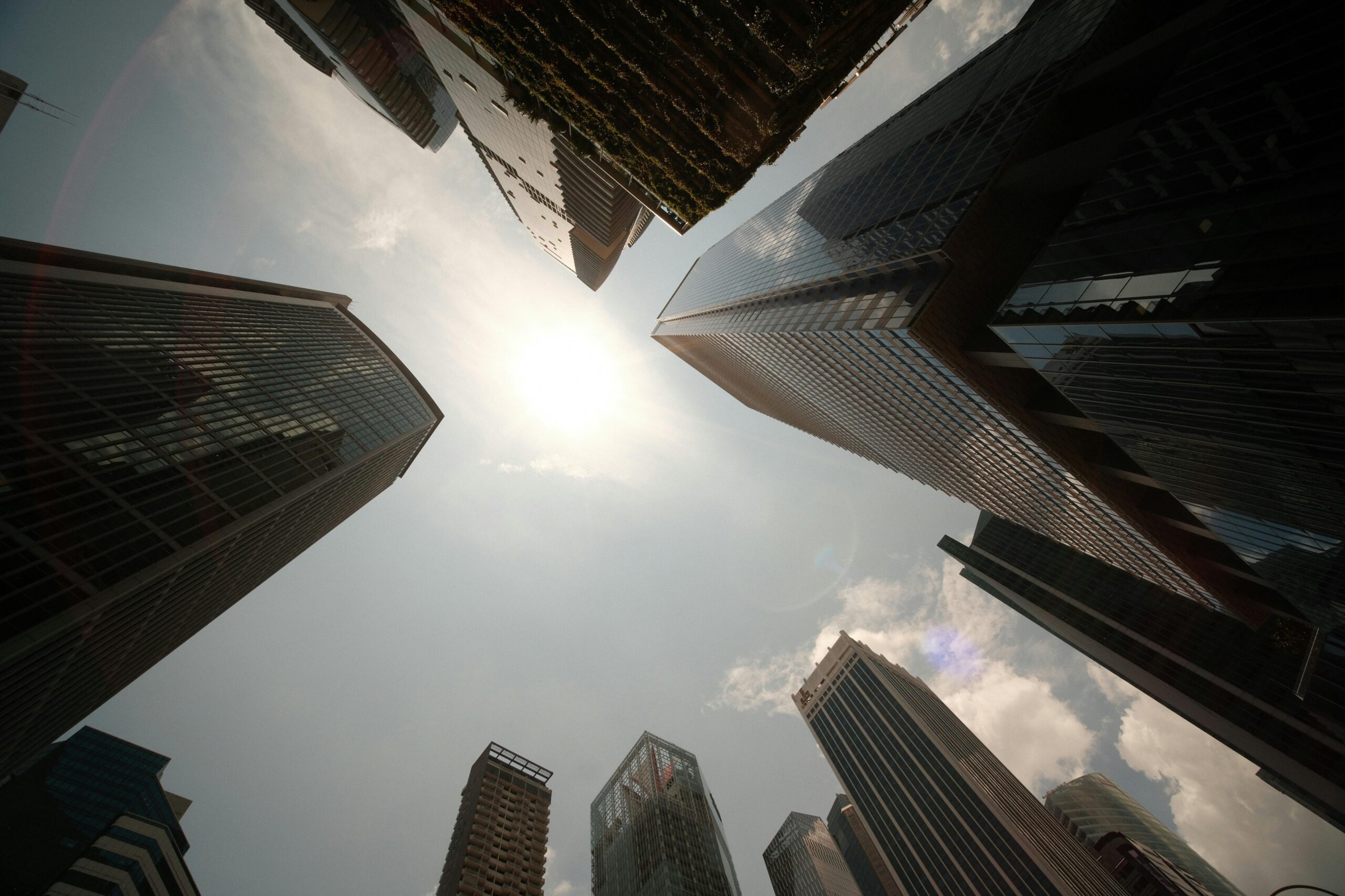 Upward view of modern skyscrapers surrounding the sun in a city skyline.