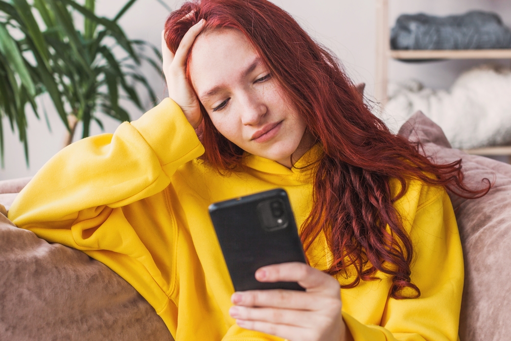 Young woman with long red hair wearing a bright yellow hoodie sits on a couch while looking at a smartphone.